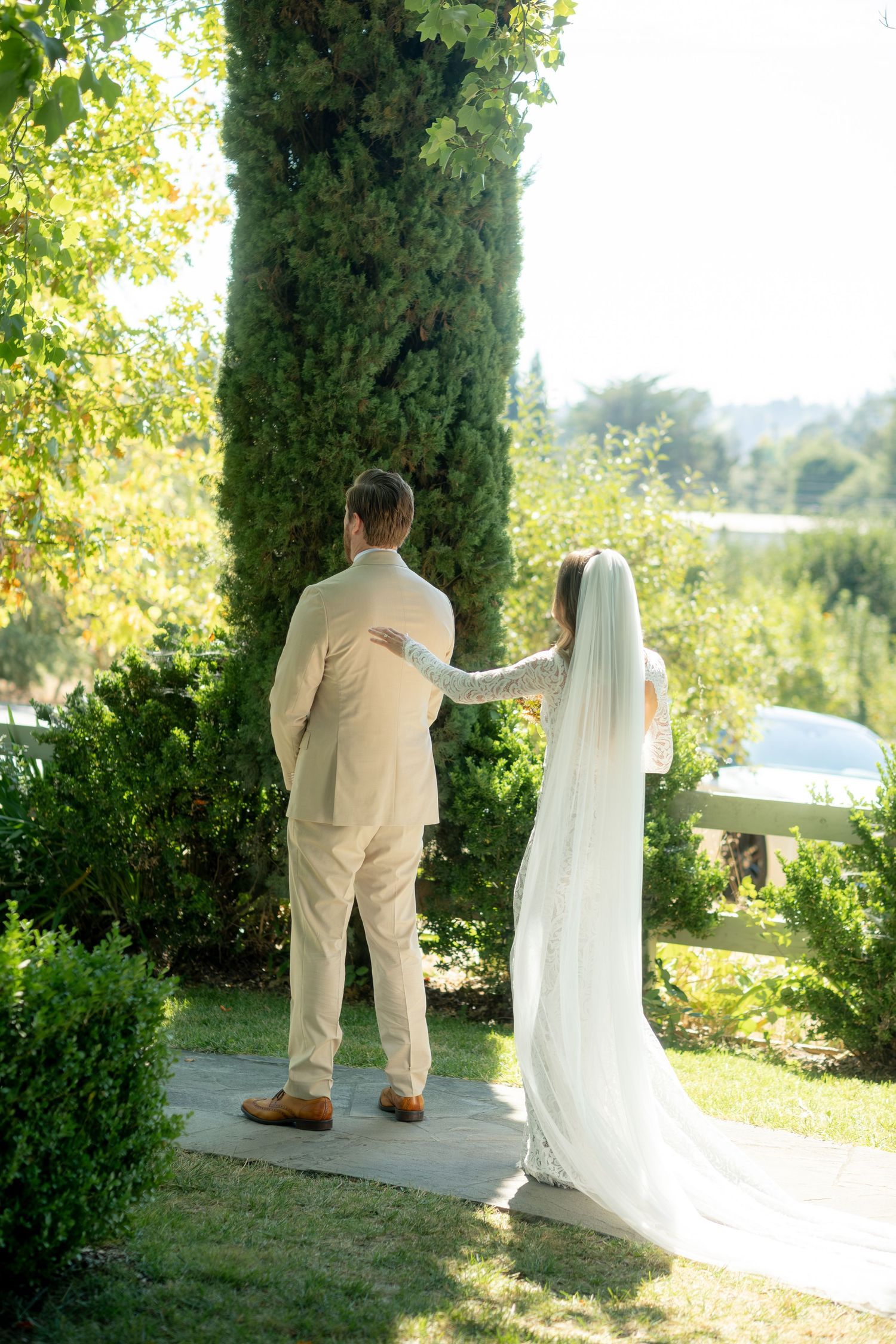 Bride and groom stand together on a garden path beside a tall cypress tree at an outdoor wedding.