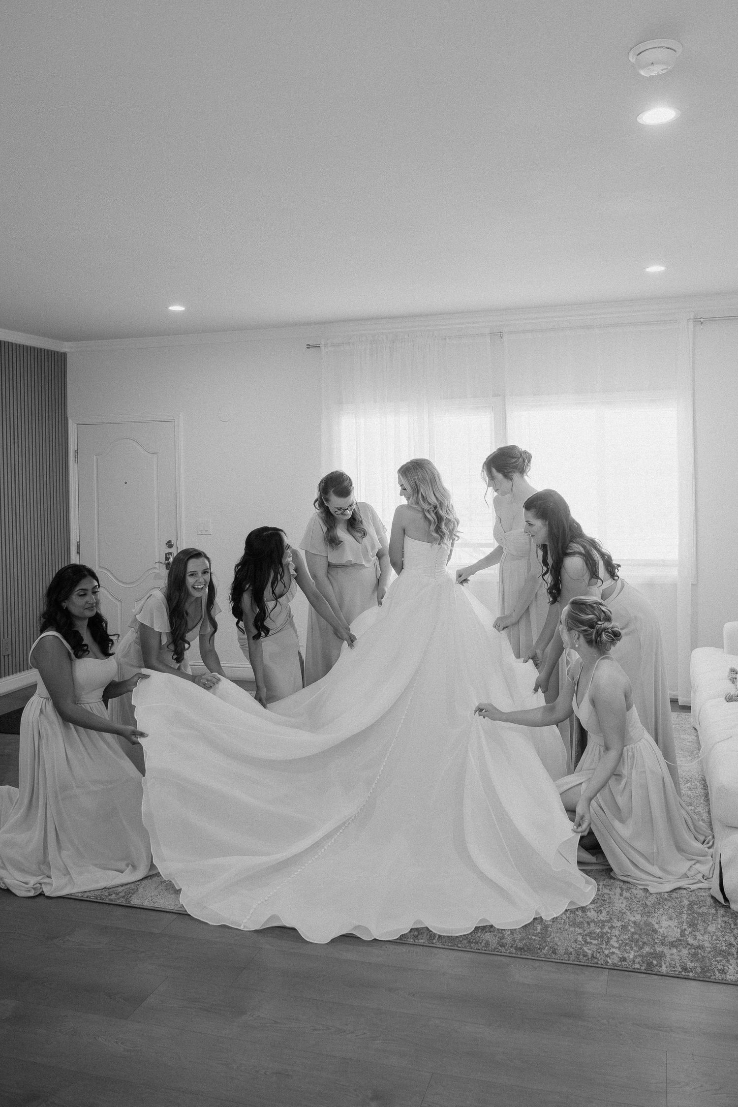 Black and white photo of bridesmaids helping bride with her wedding gown train indoors.