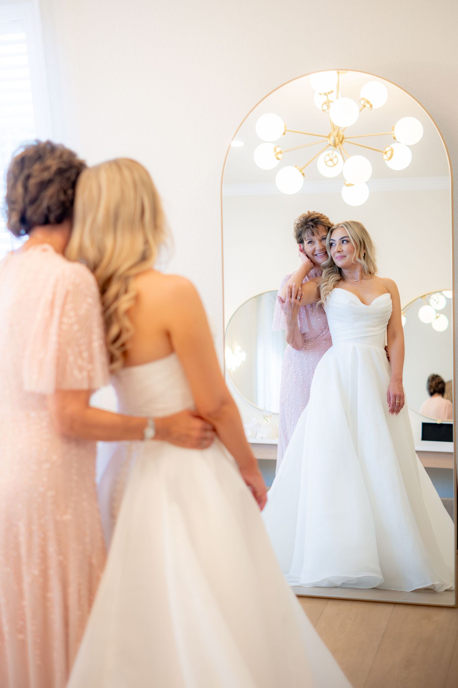 Bride in white strapless gown smiles with her mother in a mirror reflection during wedding preparations.