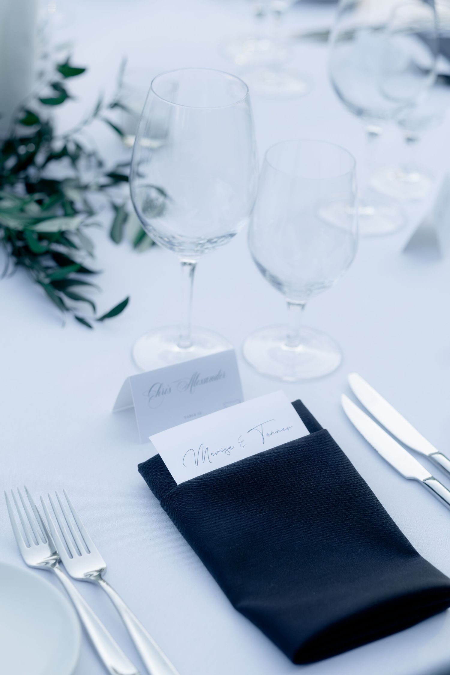 Elegant wedding place setting with black napkin, menu card, crystal glasses, and greenery.