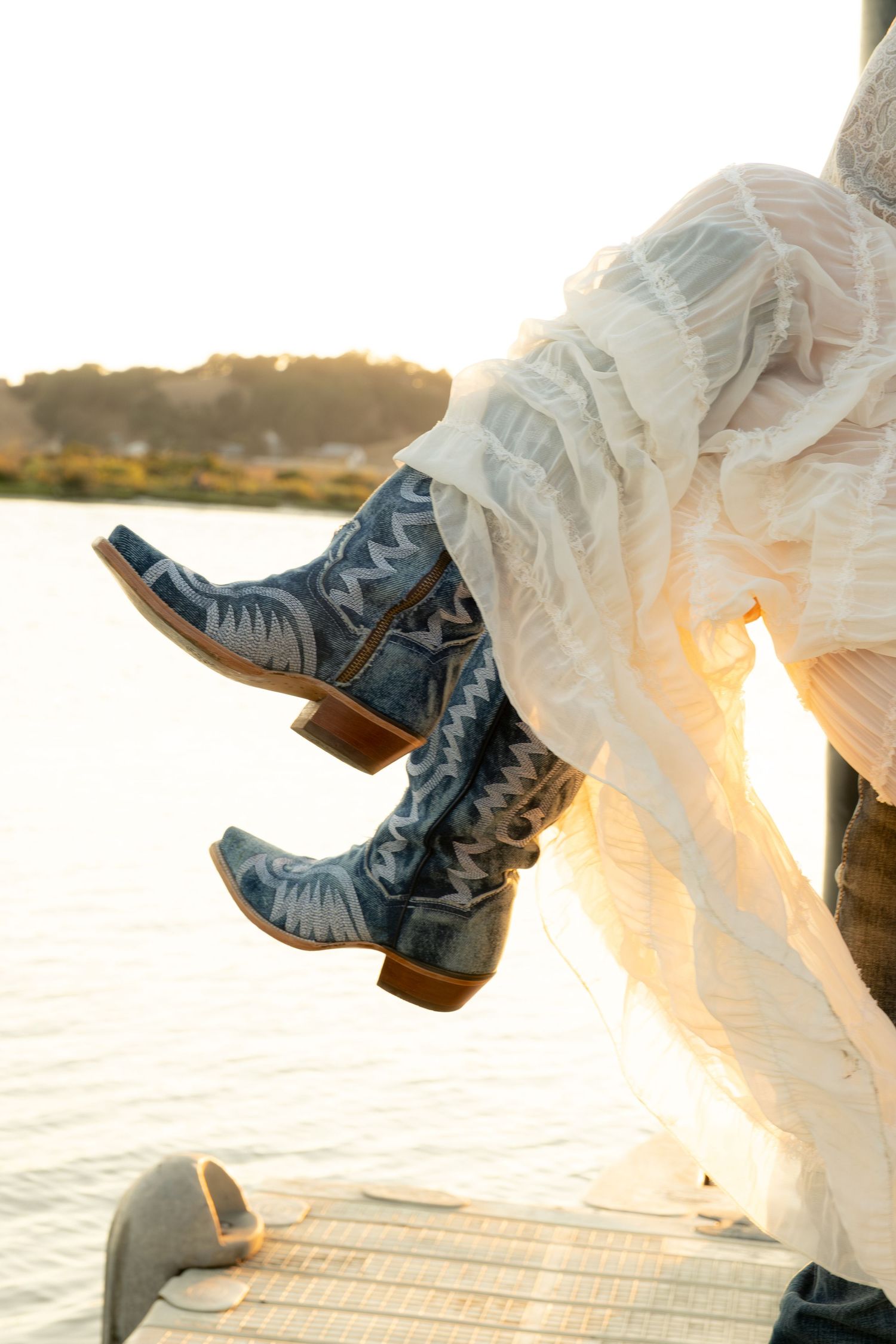 Close-up of blue embroidered cowboy boots peeking beneath a flowing white lace dress on a dock.