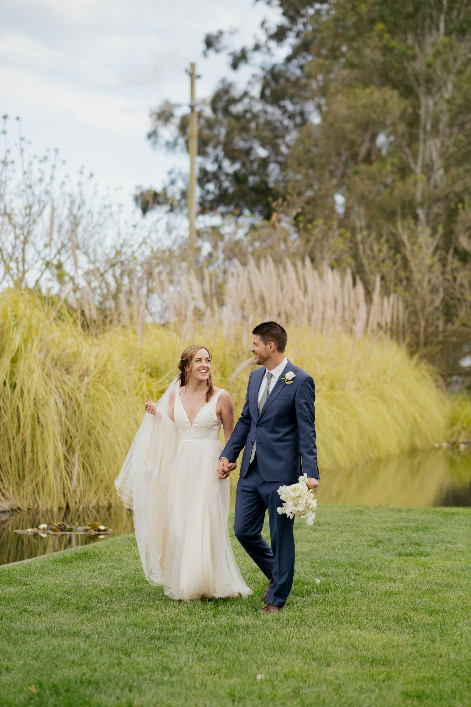 Bride and groom walk hand in hand on green lawn near a pond, bride in flowing white dress, groom in navy suit.