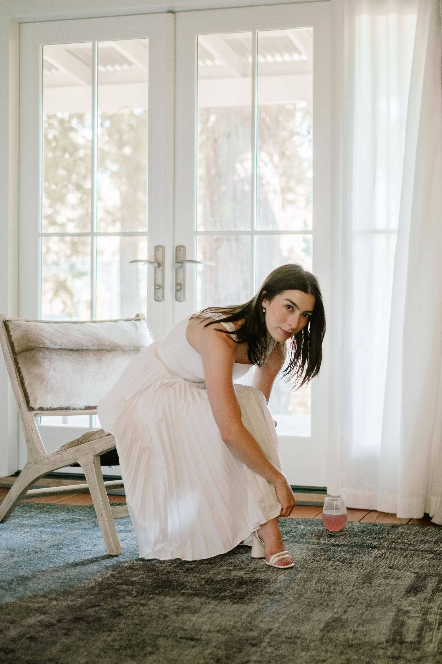 Bride in white dress adjusting her heel near French doors with soft natural light.