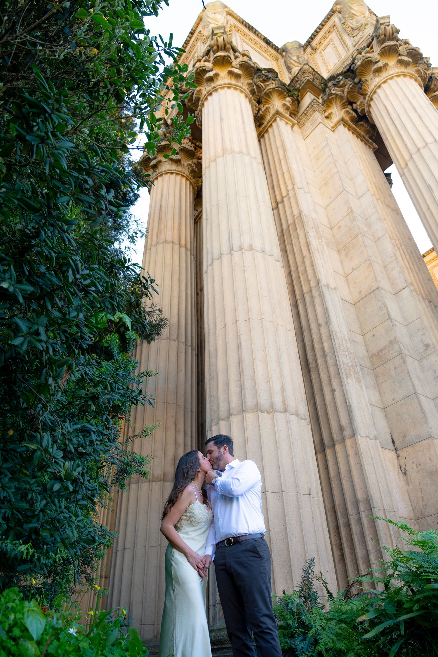 Couple sharing a romantic kiss beneath towering ornate stone columns surrounded by lush green foliage.