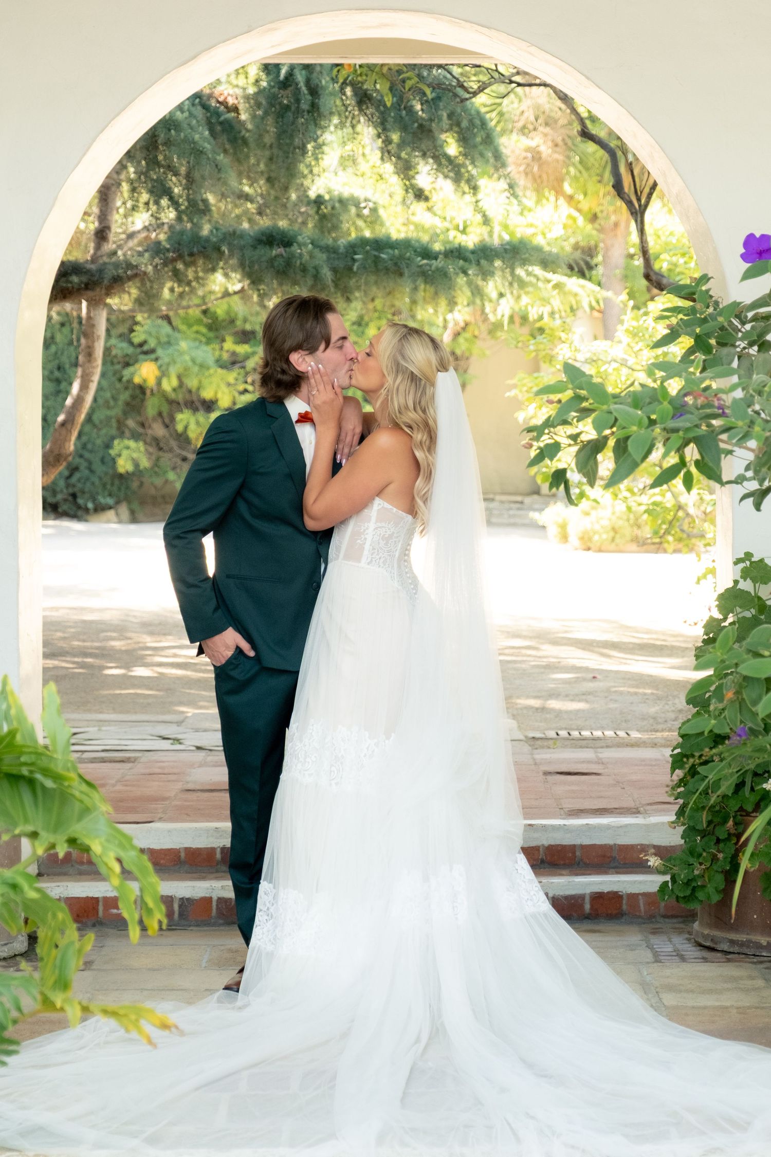 Bride and groom sharing a kiss under white arched doorway surrounded by lush garden greenery.