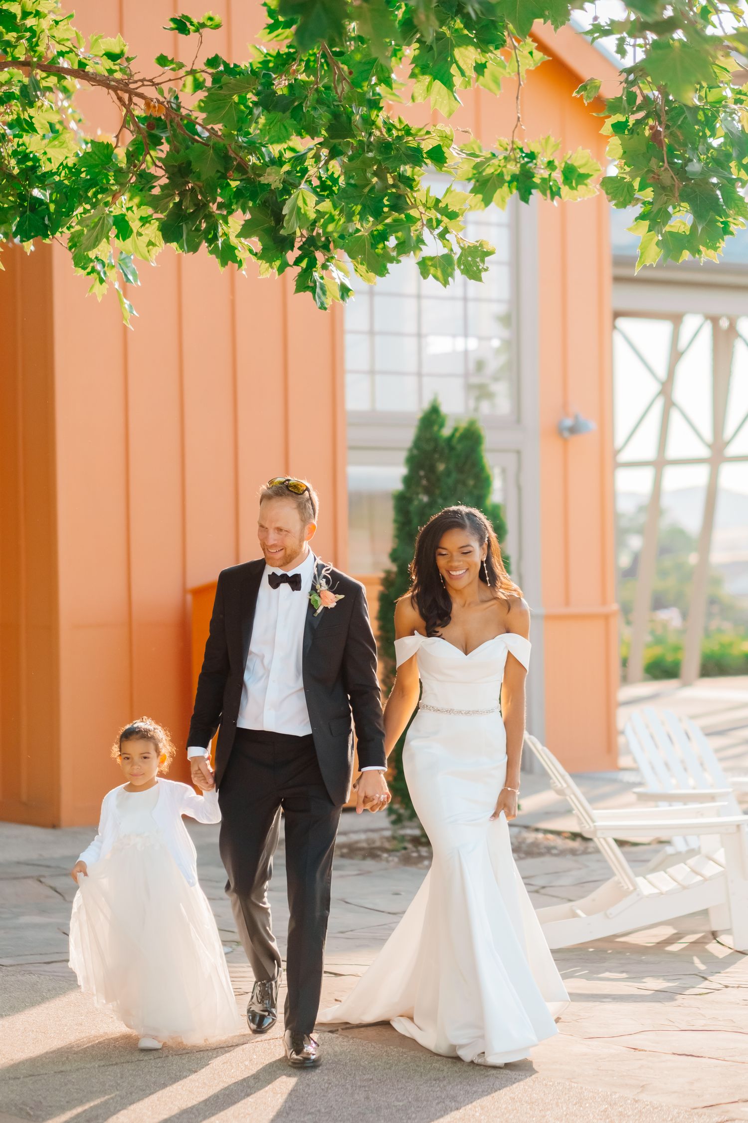 Interracial couple in wedding attire walking with flower girl beside a coral-colored building.