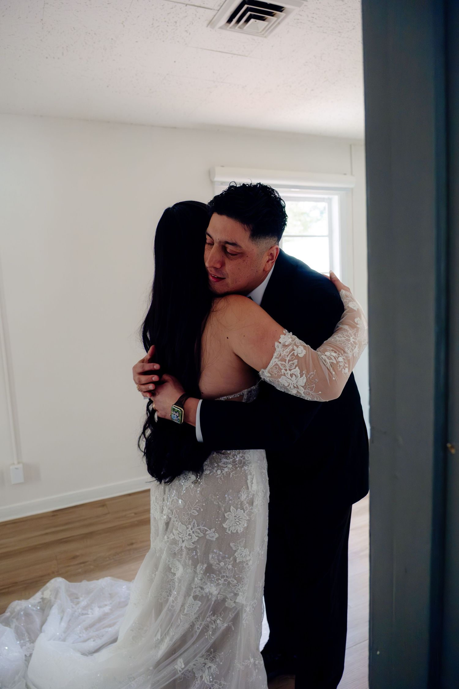 A bride in a lace gown and groom in a suit share an emotional embrace during their wedding day first look.