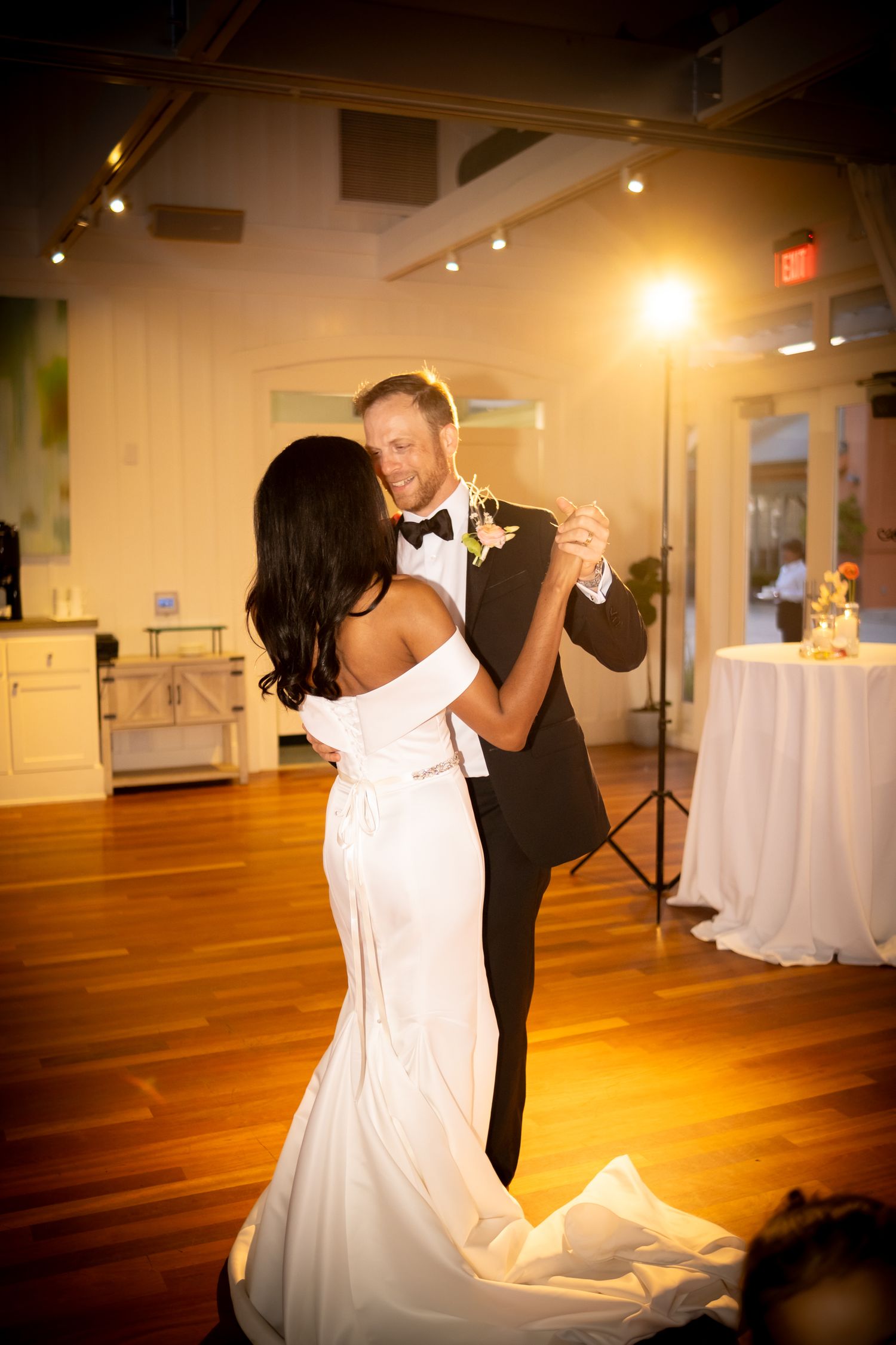 Bride in white off-shoulder gown and groom in black tuxedo share their first dance at warmly lit wedding reception.