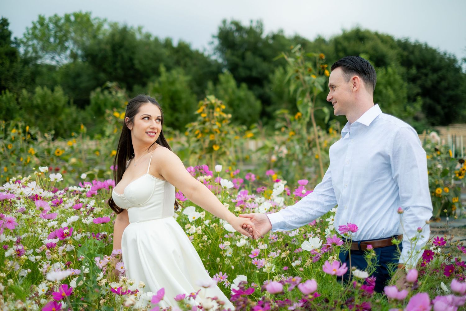 Couple holding hands in a colorful wildflower field, woman in white dress, man in blue shirt, romantic outdoor photo.