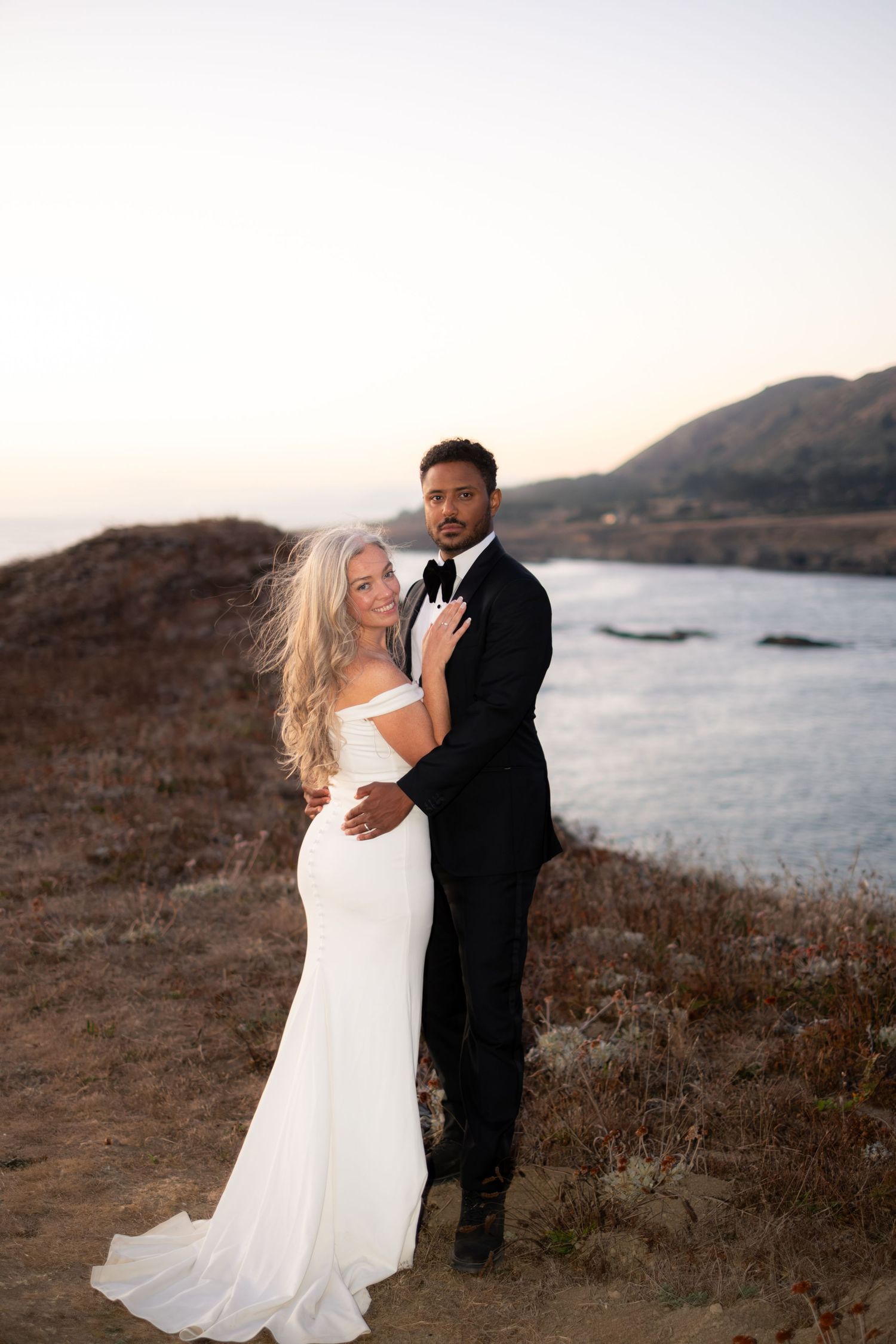 Bride in white gown and groom in black tuxedo embrace at golden hour on a coastal cliffside.