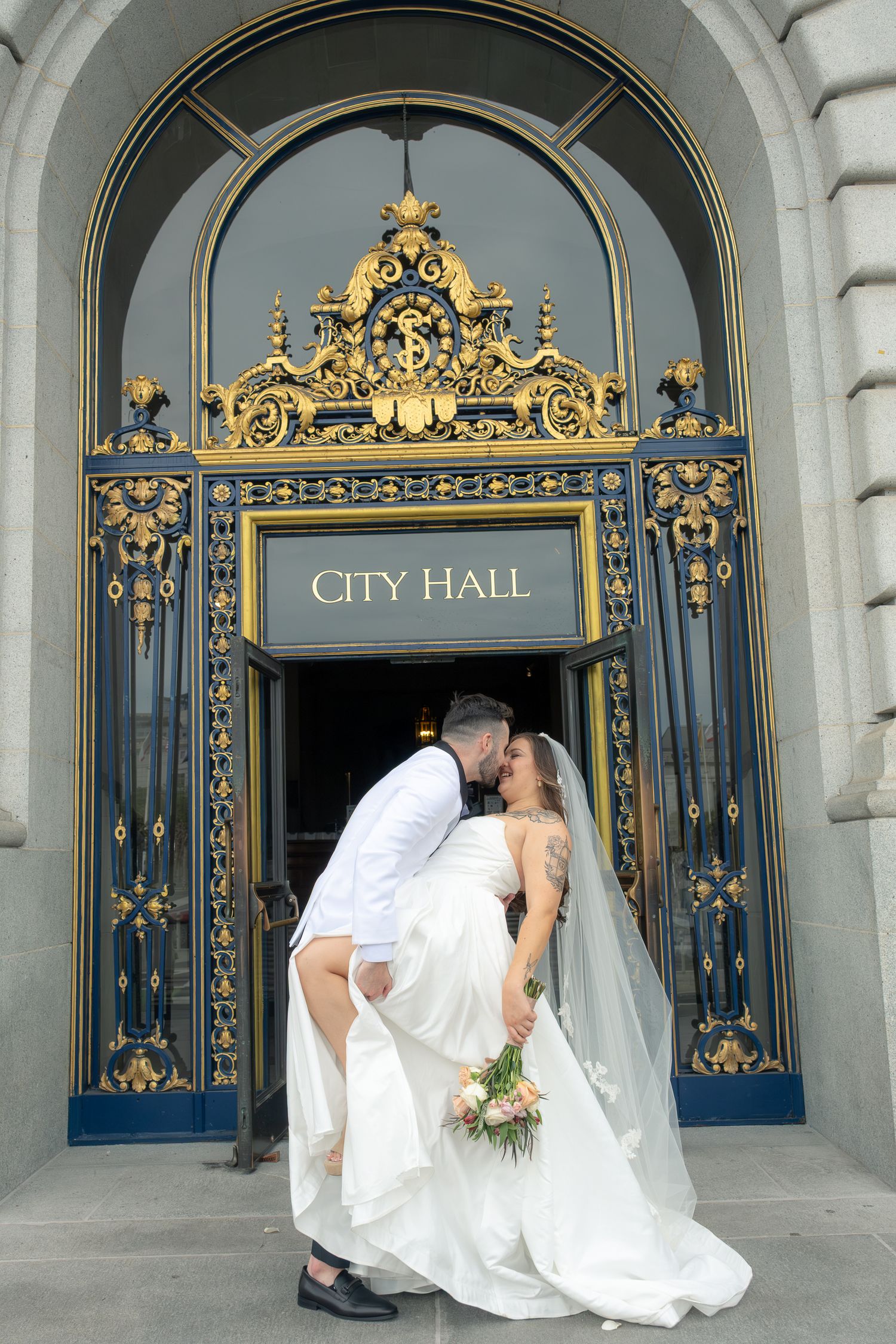 Bride and groom share a romantic dip kiss in front of San Francisco City Hall's iconic gold and blue entrance doors.