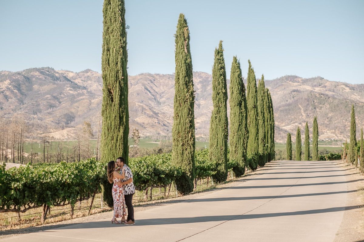 Couple kissing on a tree-lined vineyard road with mountain backdrop at a California winery.
