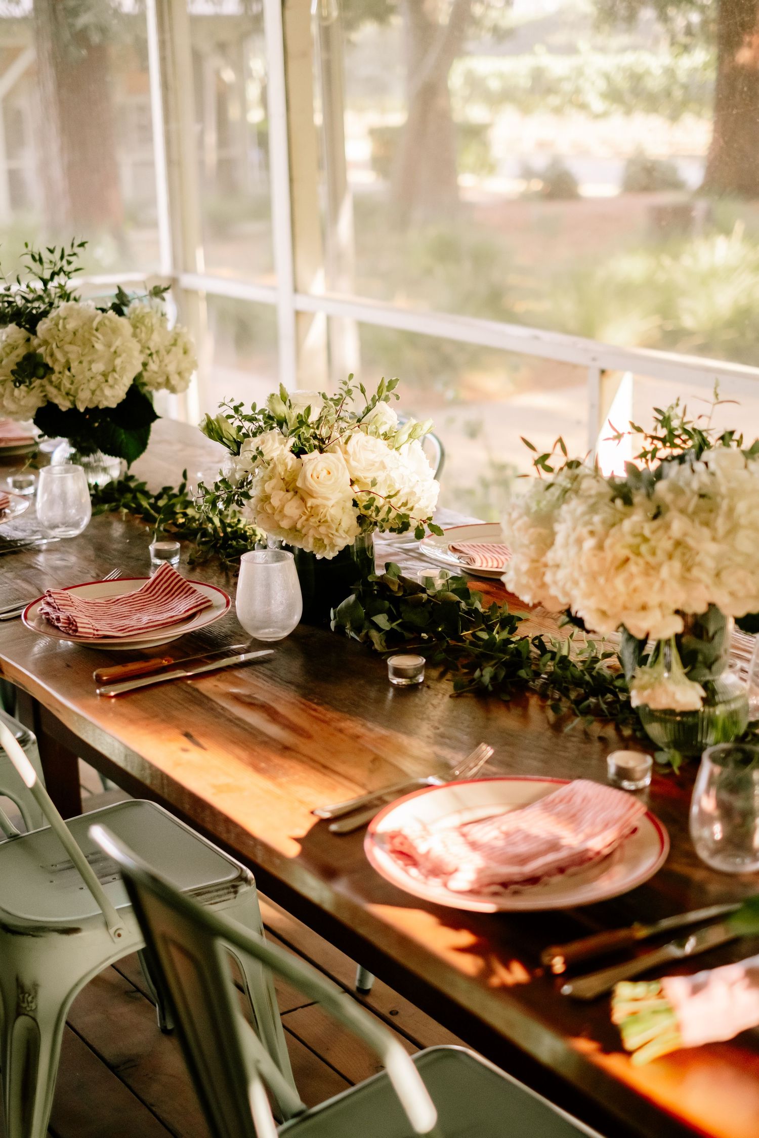 Rustic wooden dining table set with white hydrangea centerpieces and pink plates in sunlit room.
