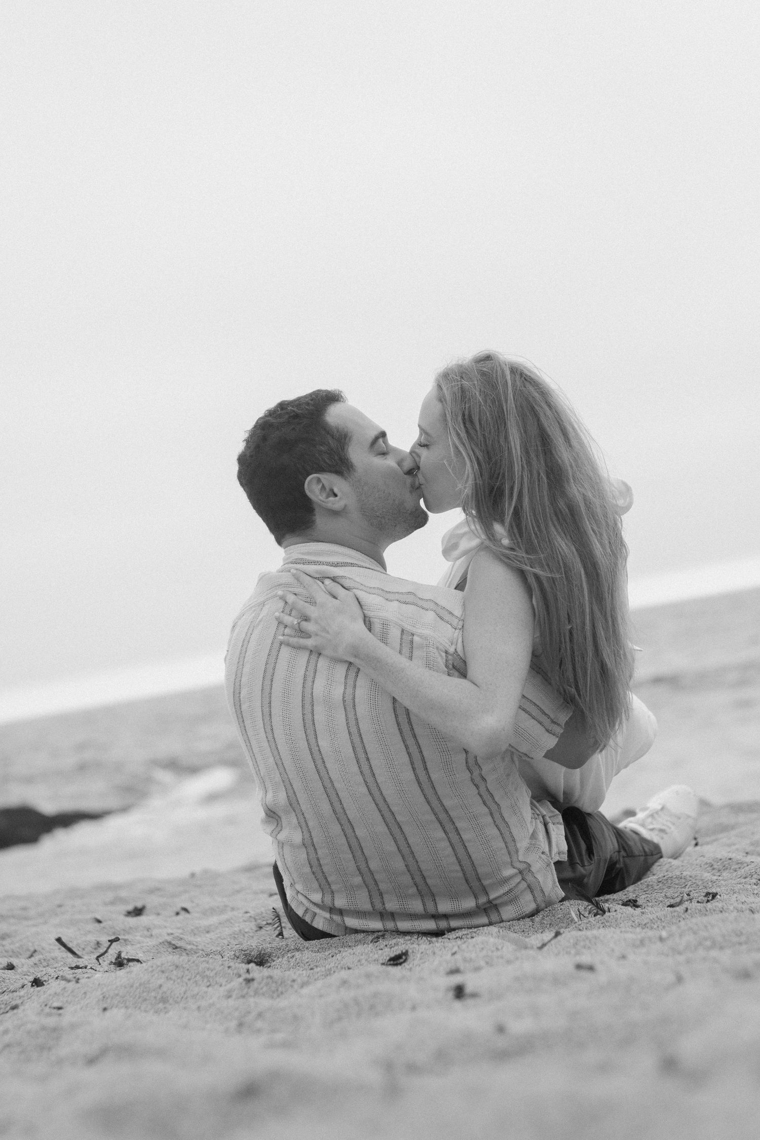 Black and white romantic photo of couple kissing while sitting together on sandy beach by ocean.