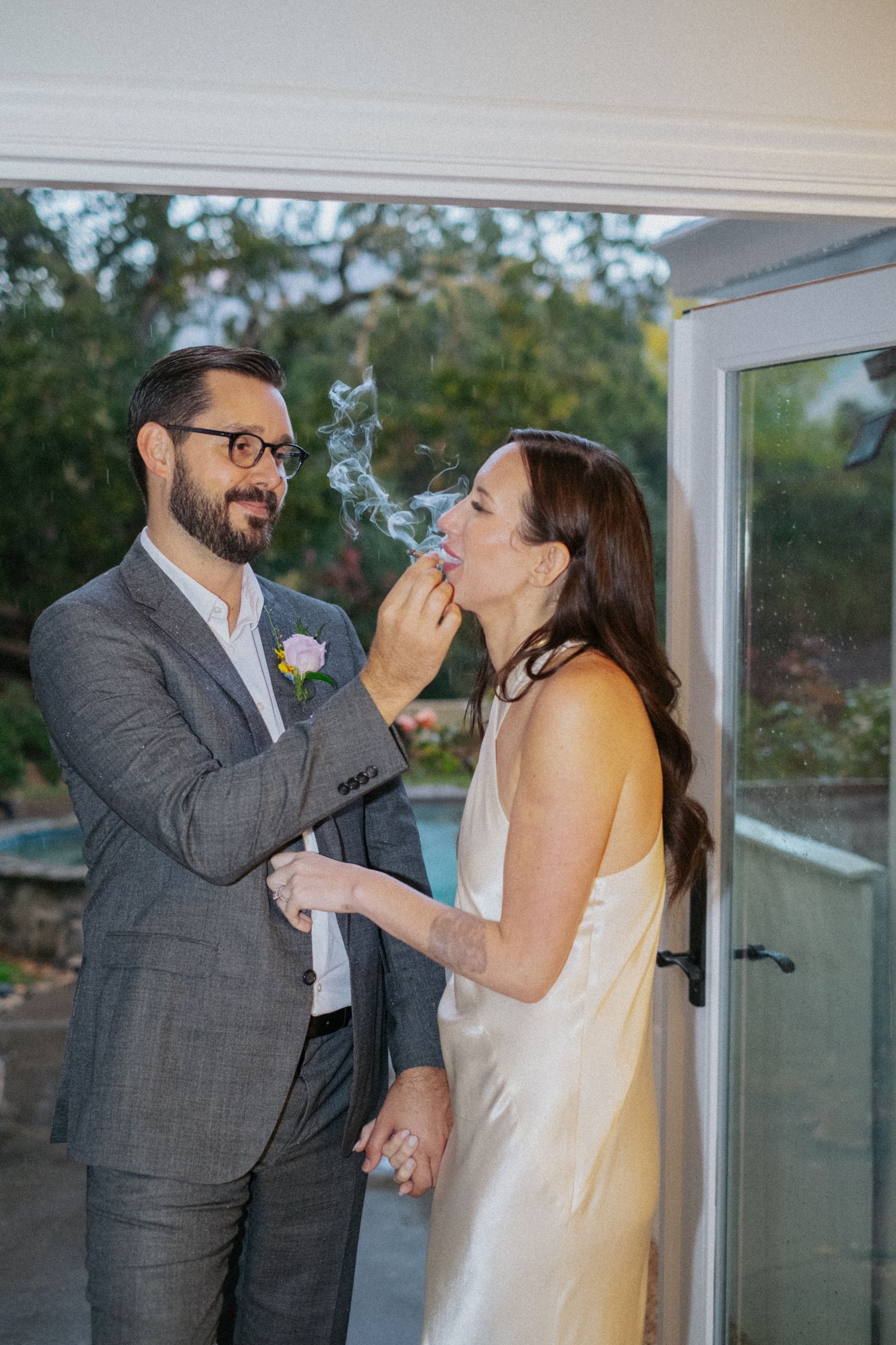 Bride in white satin dress shares a romantic moment with groom in gray suit while smoking near a glass door.
