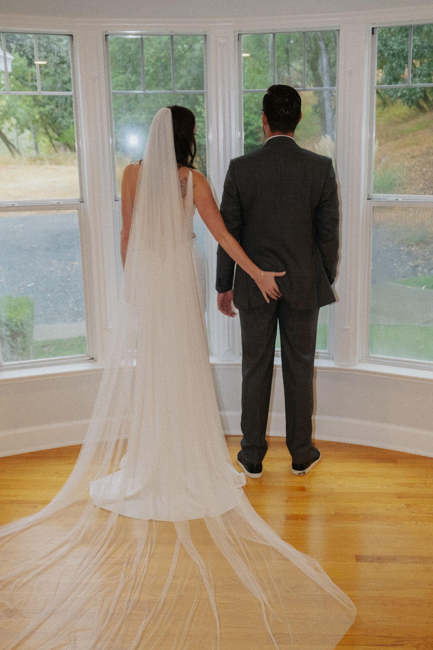 Bride and groom stand hand-in-hand before bay windows, her cathedral veil trailing on hardwood floors.