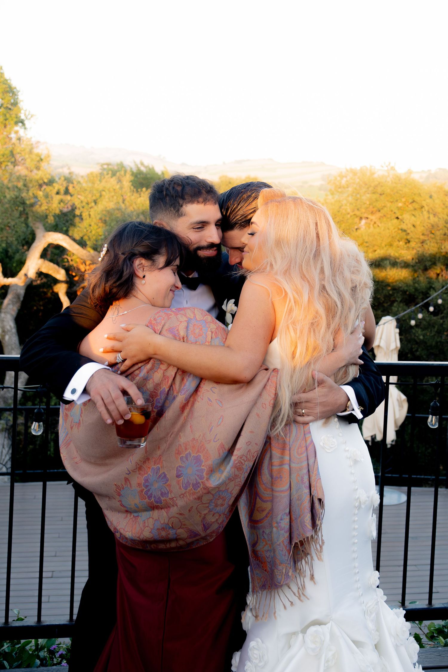 Joyful newlyweds share a warm group hug with a female guest on an outdoor balcony at golden hour.