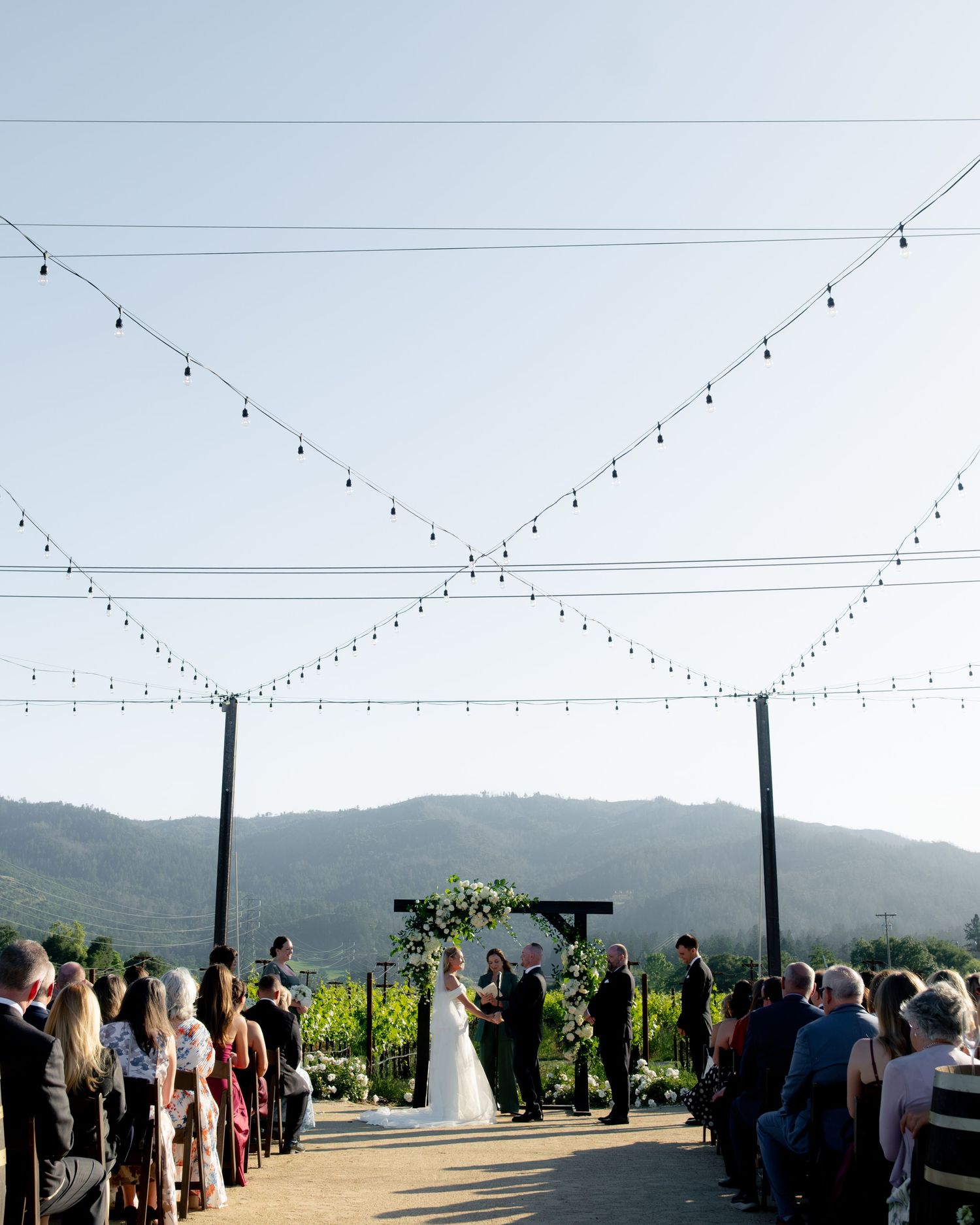 Outdoor wedding ceremony with string lights, floral arch, mountain backdrop, and seated guests.