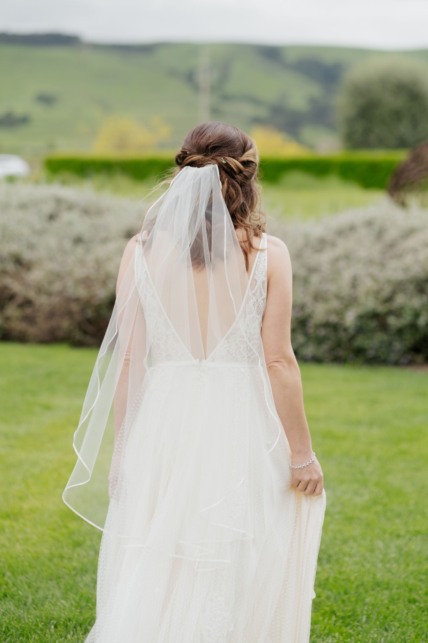 Bride in a boho tiered white gown with veil and braided updo walks through a lush green garden landscape.