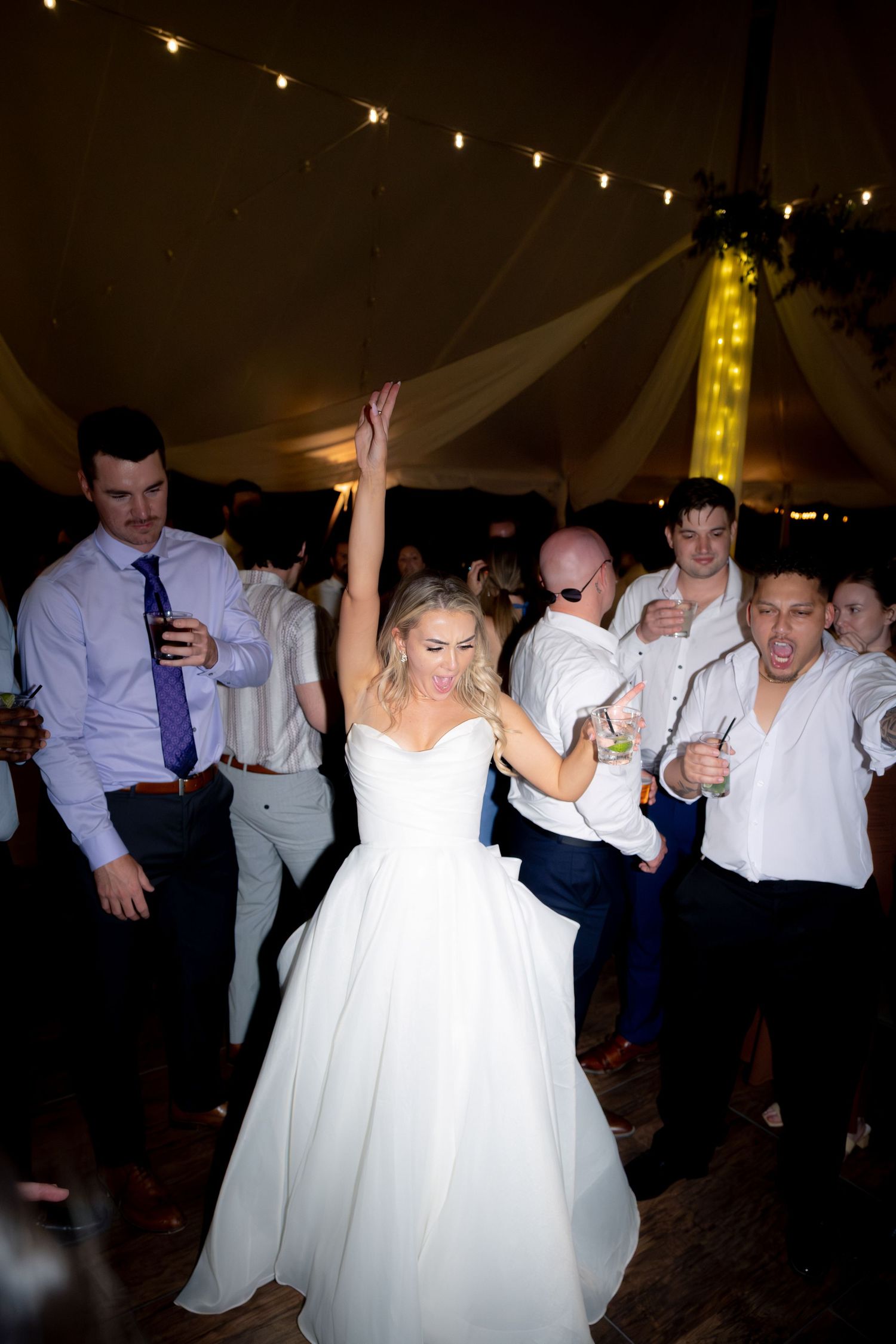 Joyful bride in white gown raises her hand while dancing with guests at wedding reception.
