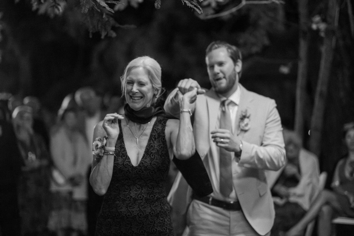 Groom escorts smiling woman down the aisle at an outdoor wedding ceremony in black and white photo.