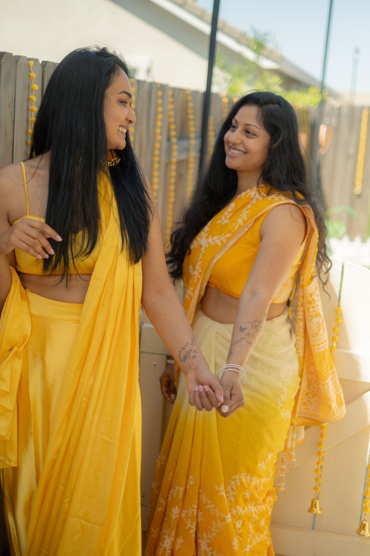 Two women in matching yellow sarees smile and hold hands at an Indian celebration.