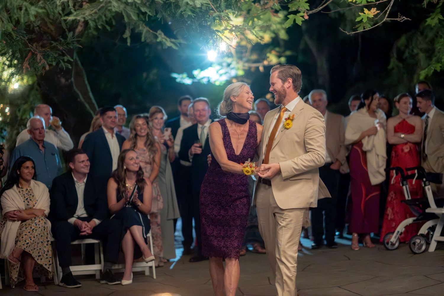 Groom dances with a woman in purple dress at an evening outdoor wedding reception with guests watching.
