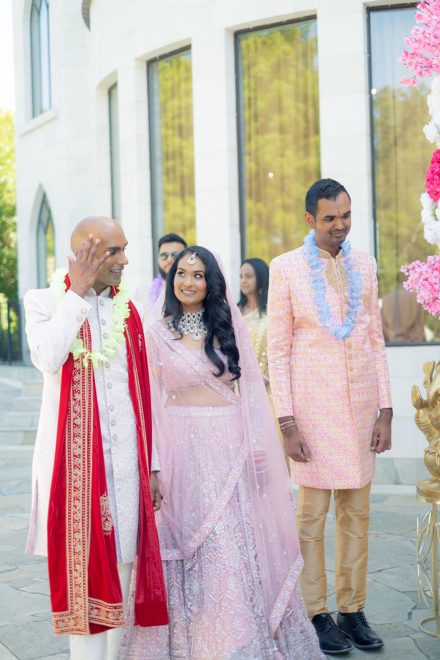 Indian bride in pink lehenga stands with emotional father and groom during outdoor wedding ceremony.