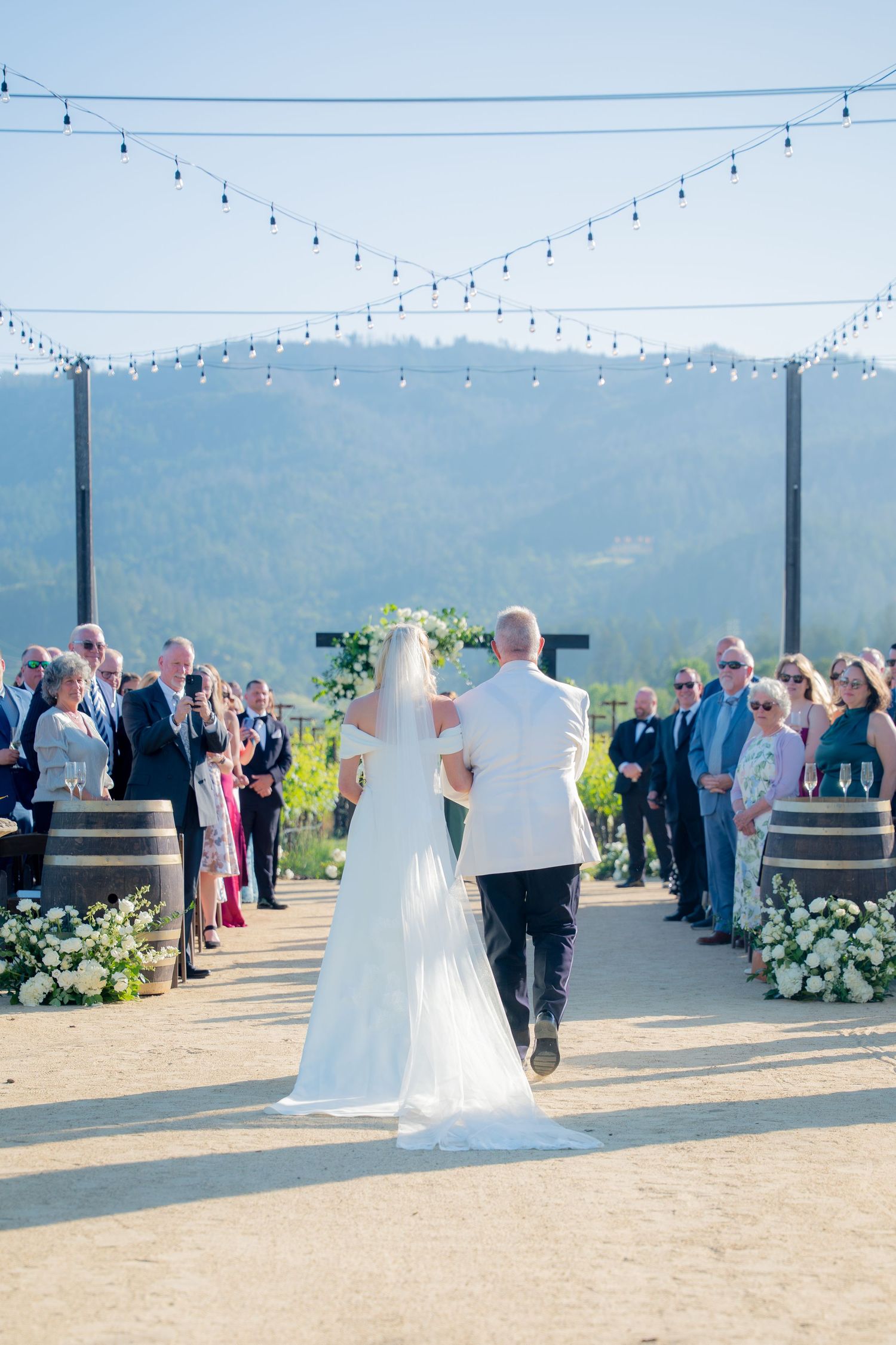 Bride and escort walk down outdoor wedding aisle with mountain views and string lights overhead.