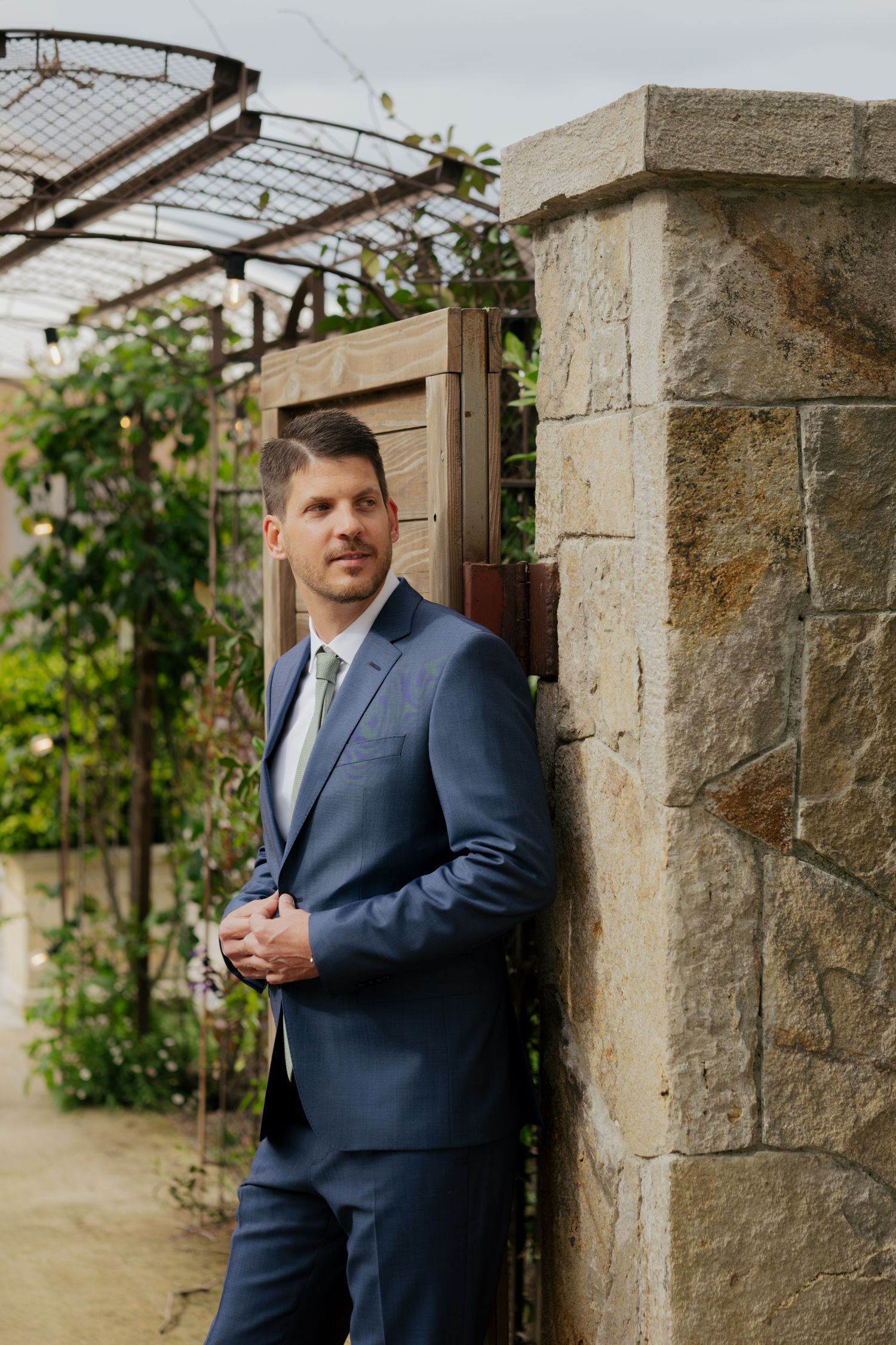 Handsome groom in navy blue suit leans against a stone pillar beneath a vine-covered pergola at a garden wedding venue.