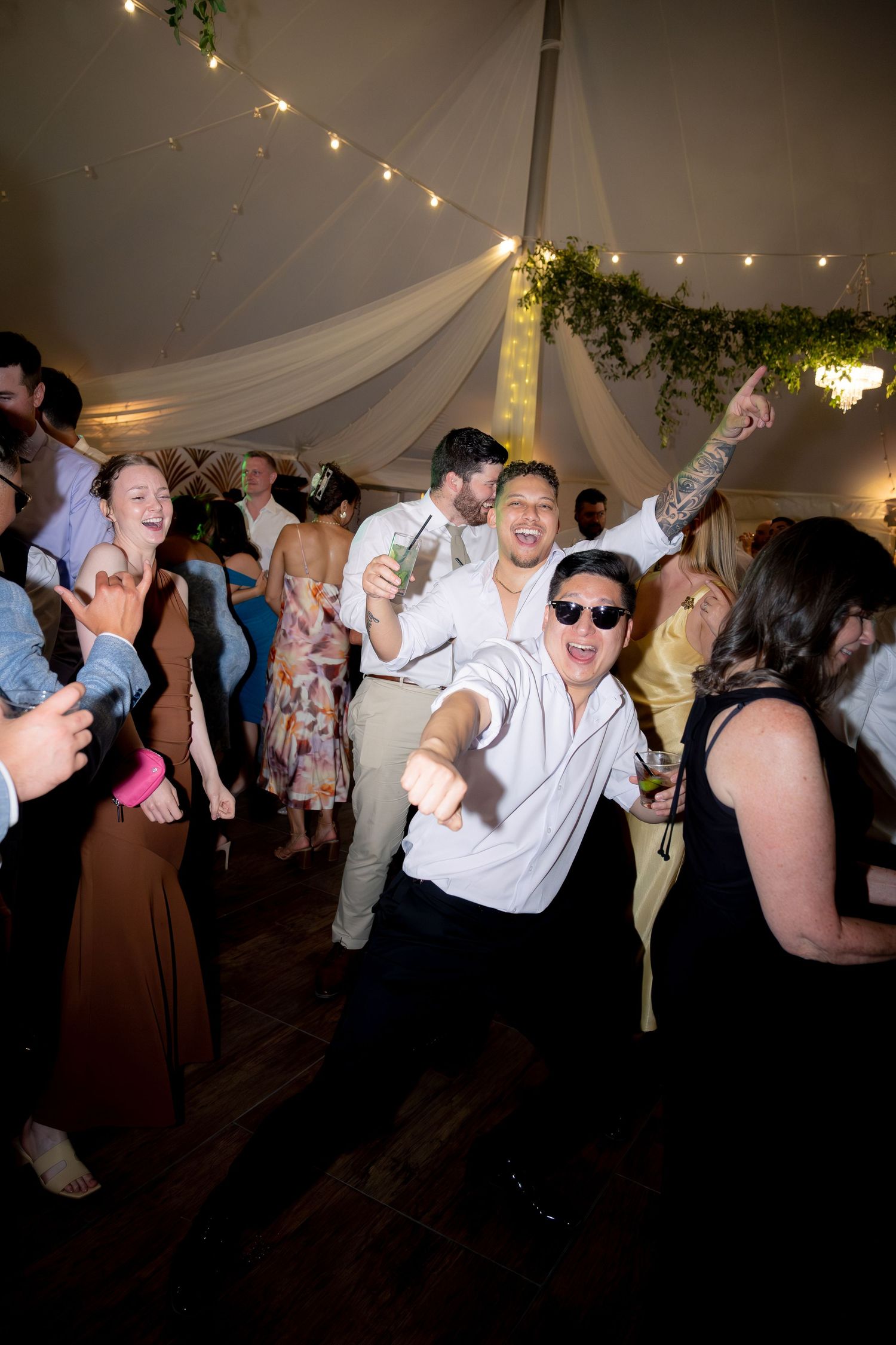 Energetic wedding guests dancing and celebrating under string lights in a decorated reception tent.