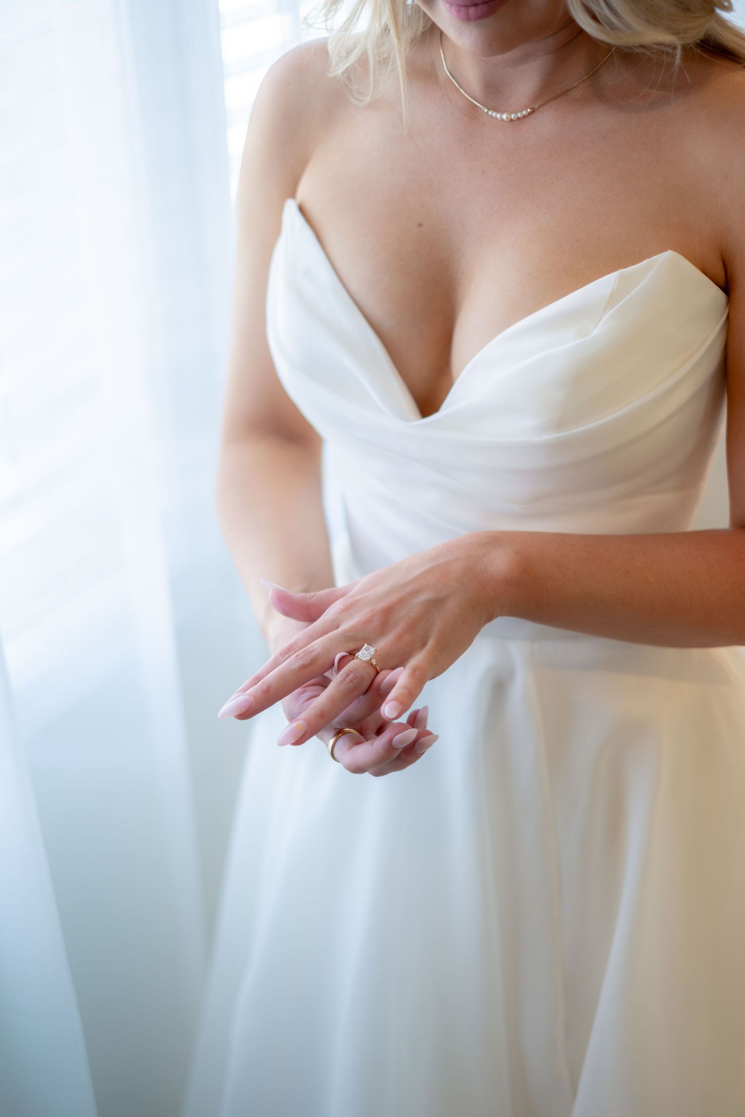 Bride in strapless white gown admiring engagement ring by window.