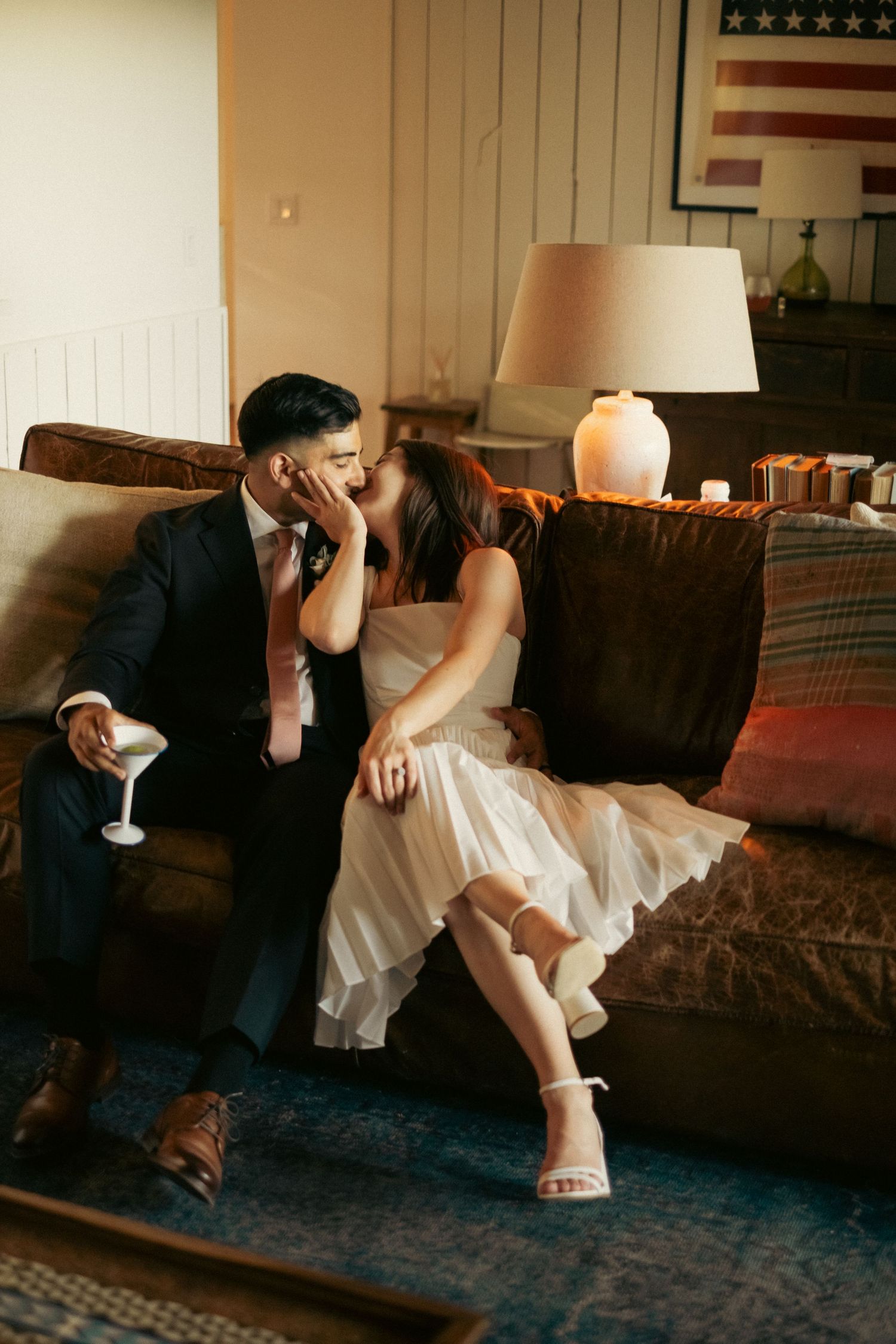 Bride and groom share a kiss while seated on a couch in warmly lit rustic interior.