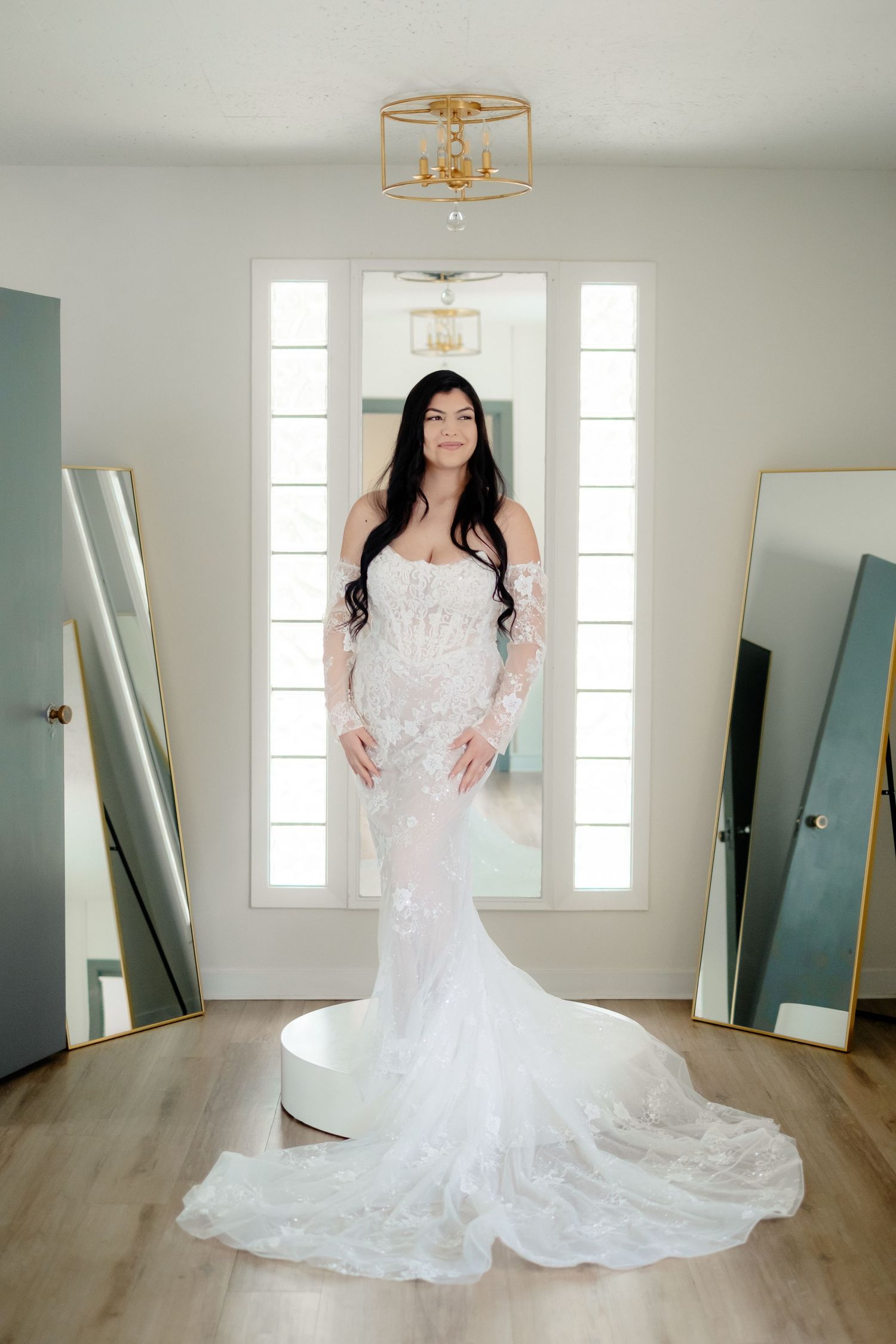 Bride in a fitted white lace mermaid wedding gown with long sleeves posing in a bridal suite.