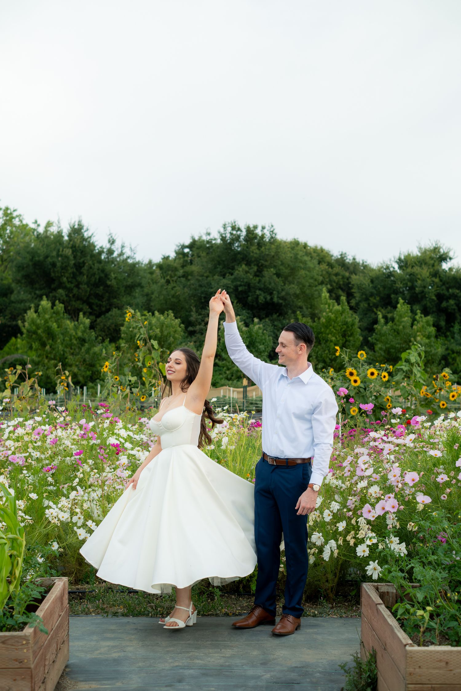 Bride and groom dancing in a vibrant flower garden, man spinning woman in white tea-length wedding dress.