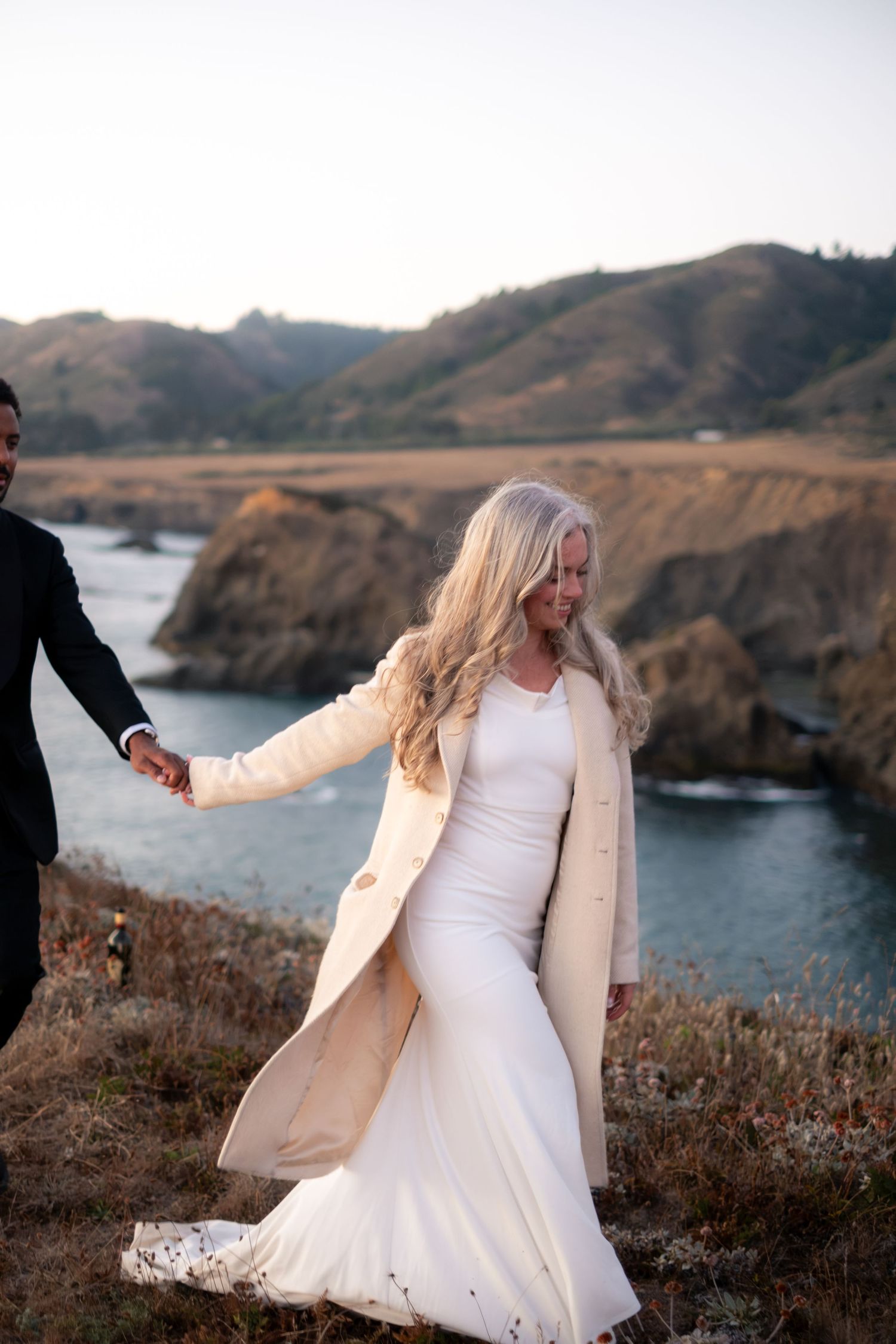 Blonde bride in white gown and cream coat holding hands with groom on coastal cliffside.