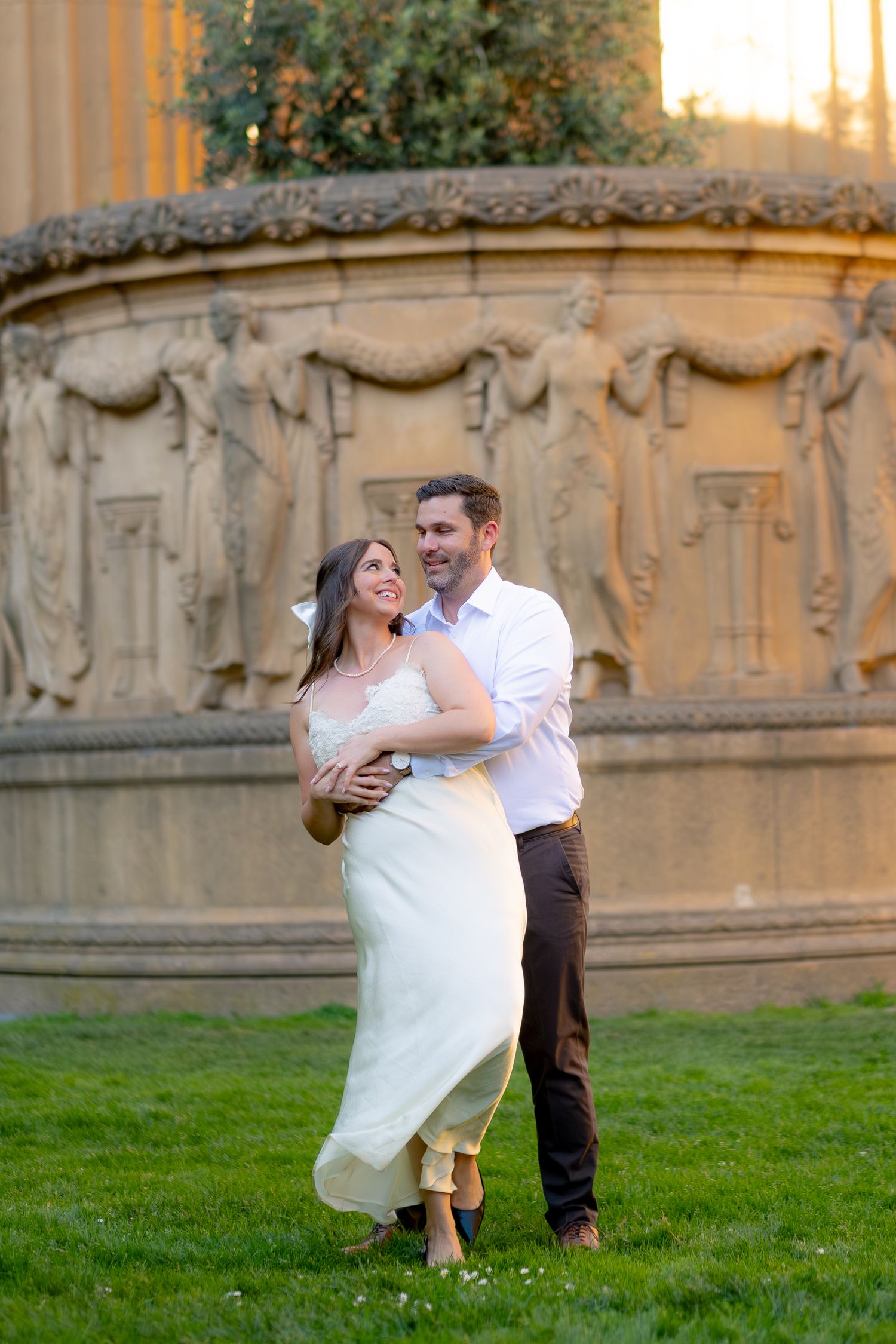 Couple embracing and smiling on green grass in front of ornate stone fountain at Palace of Fine Arts.
