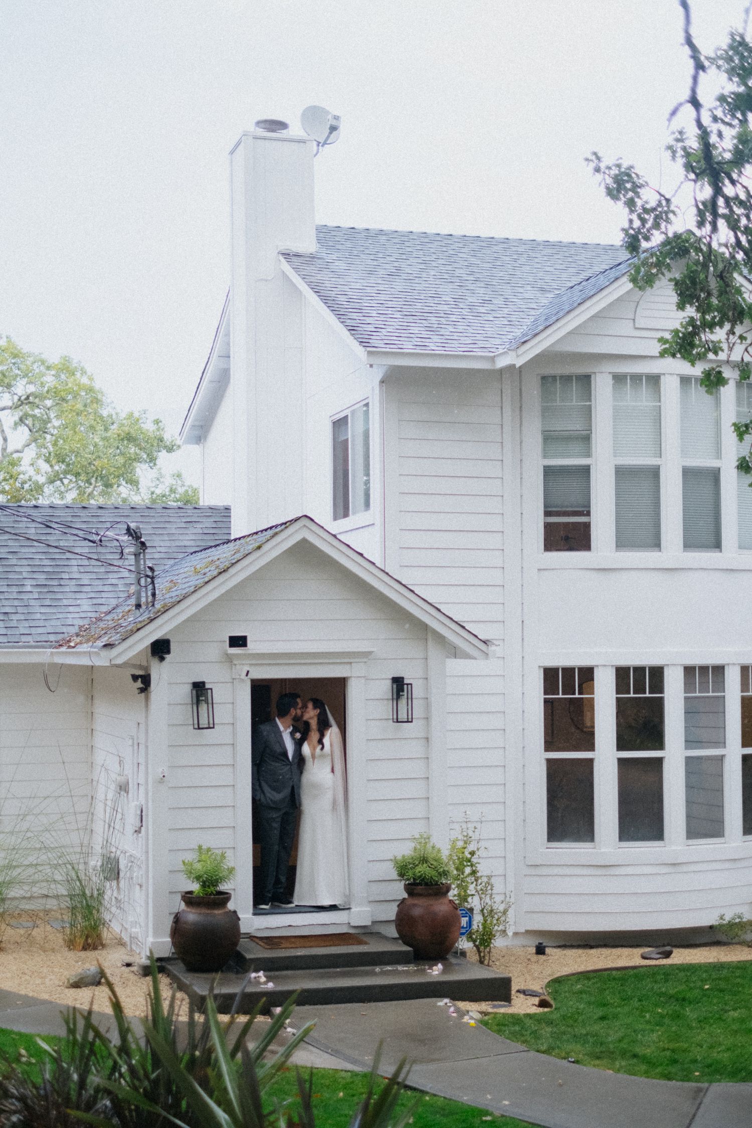 A bride and groom share a kiss in the doorway of a charming white two-story farmhouse wedding venue.