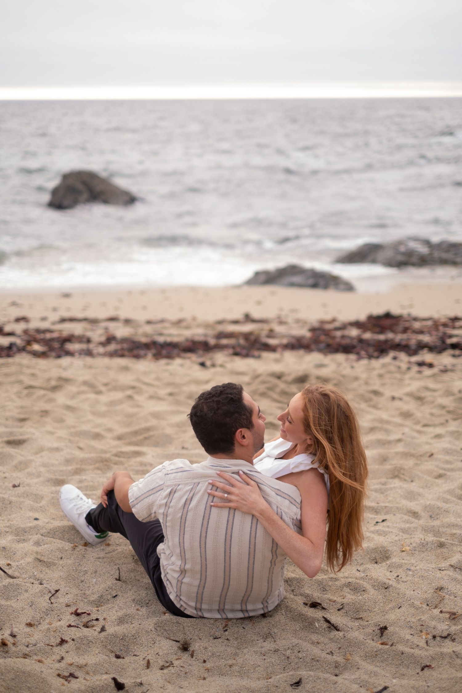 Couple sitting together on sandy beach gazing at each other with ocean in background.