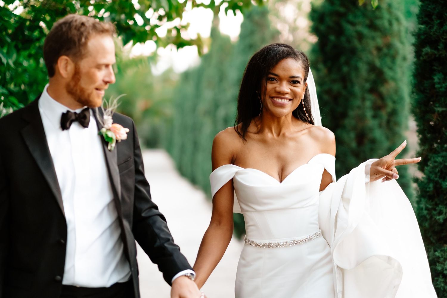 Smiling bride in off-shoulder gown holds hands with groom in black tuxedo while walking outdoors.