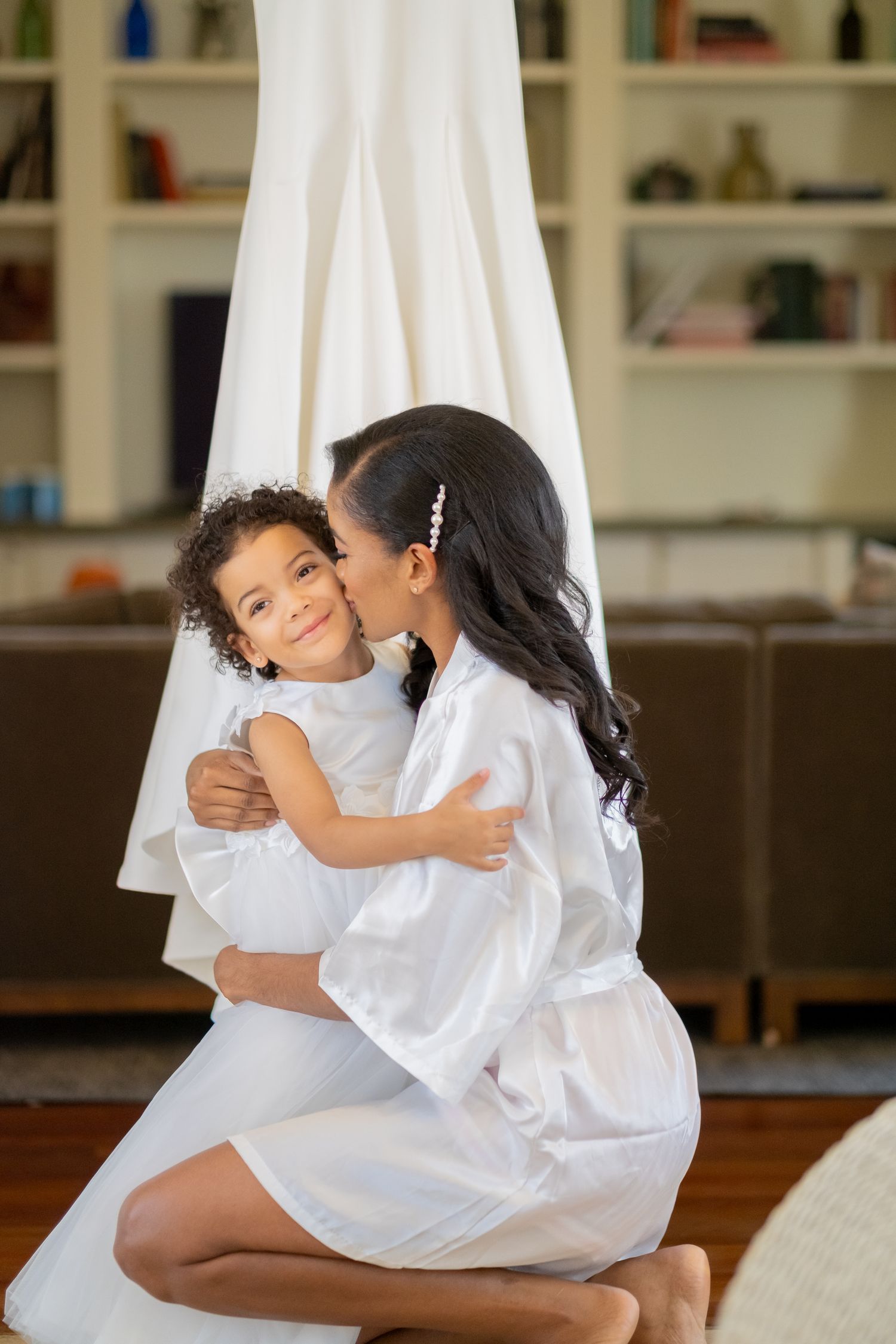 Bride in white robe kissing young flower girl in white dress during wedding preparations at home.