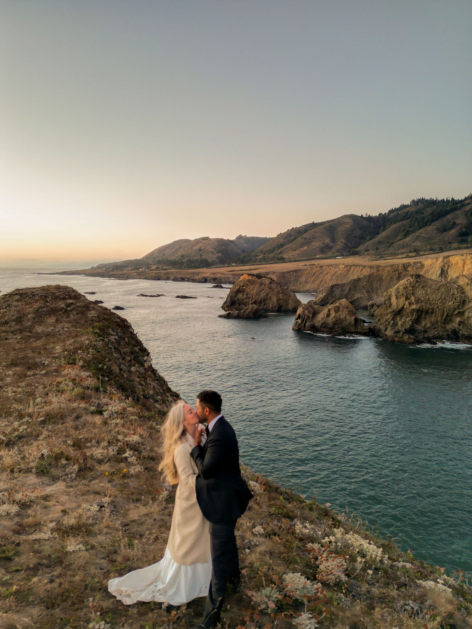 Bride and groom sharing a romantic kiss on a coastal cliff at sunset with stunning ocean and mountain views.