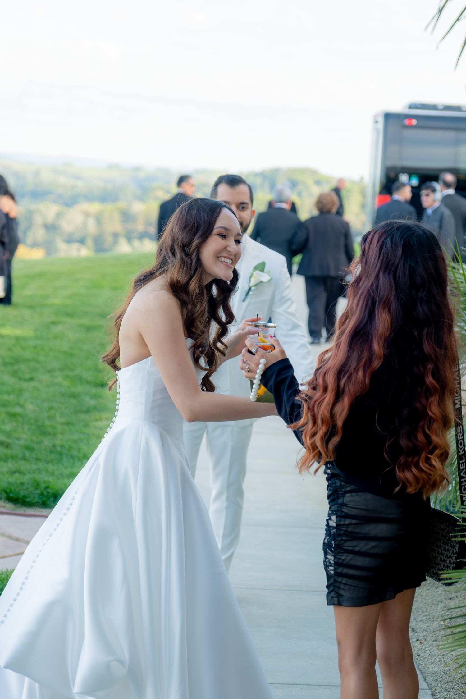 Bride in white strapless gown greeting a guest in black dress at outdoor wedding reception.