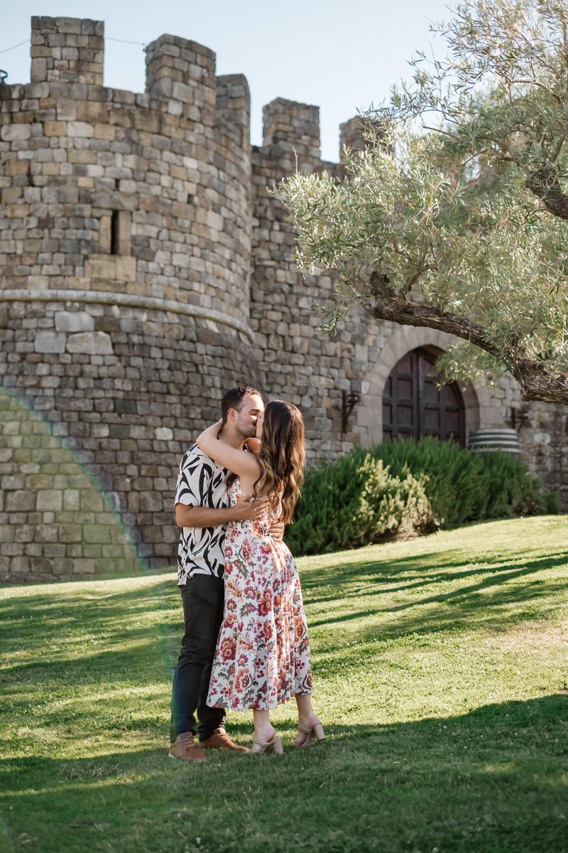Couple embracing and kissing on green lawn in front of a stone castle building.