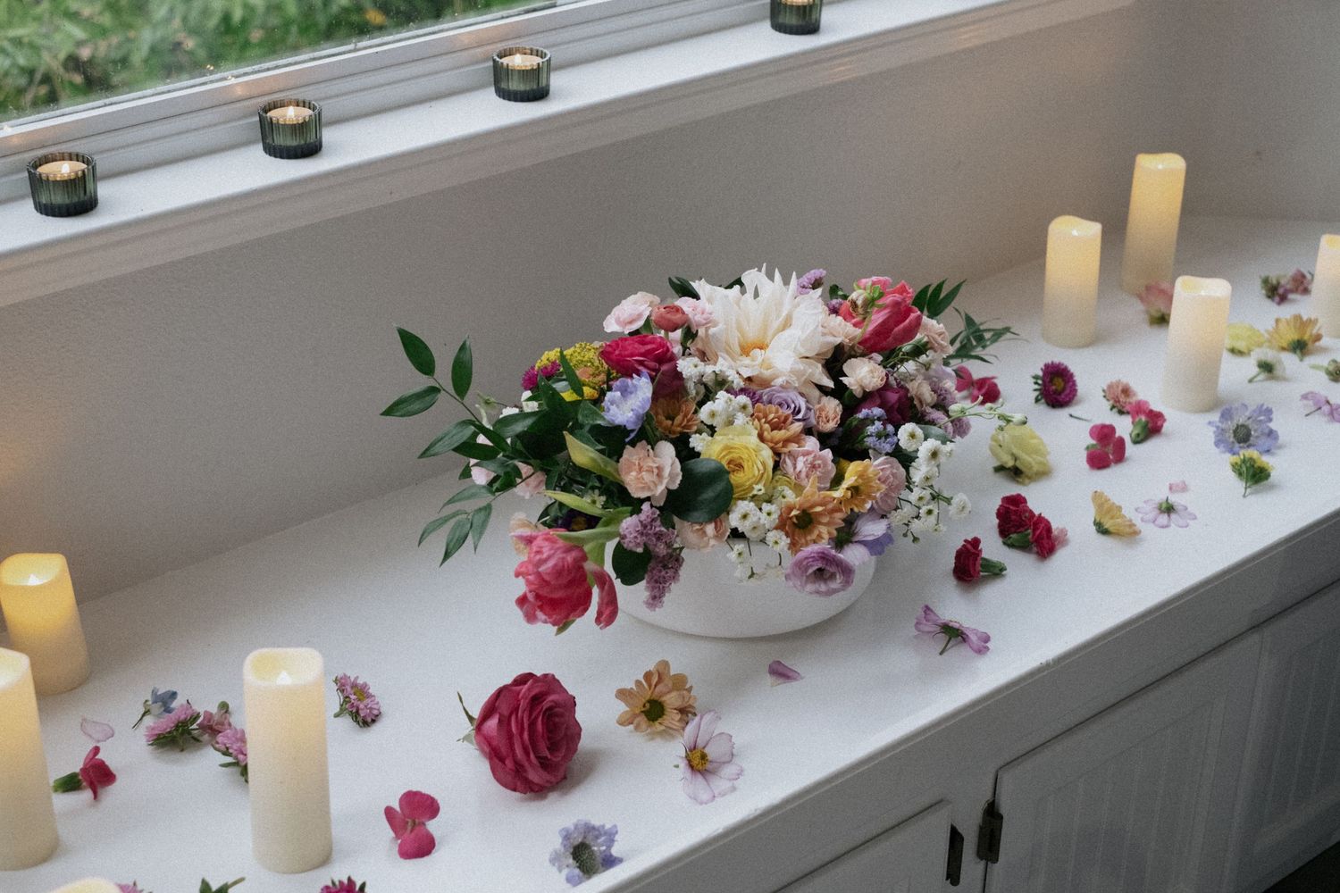 Colorful wedding floral arrangement with roses and mixed blooms surrounded by pillar candles on a windowsill.