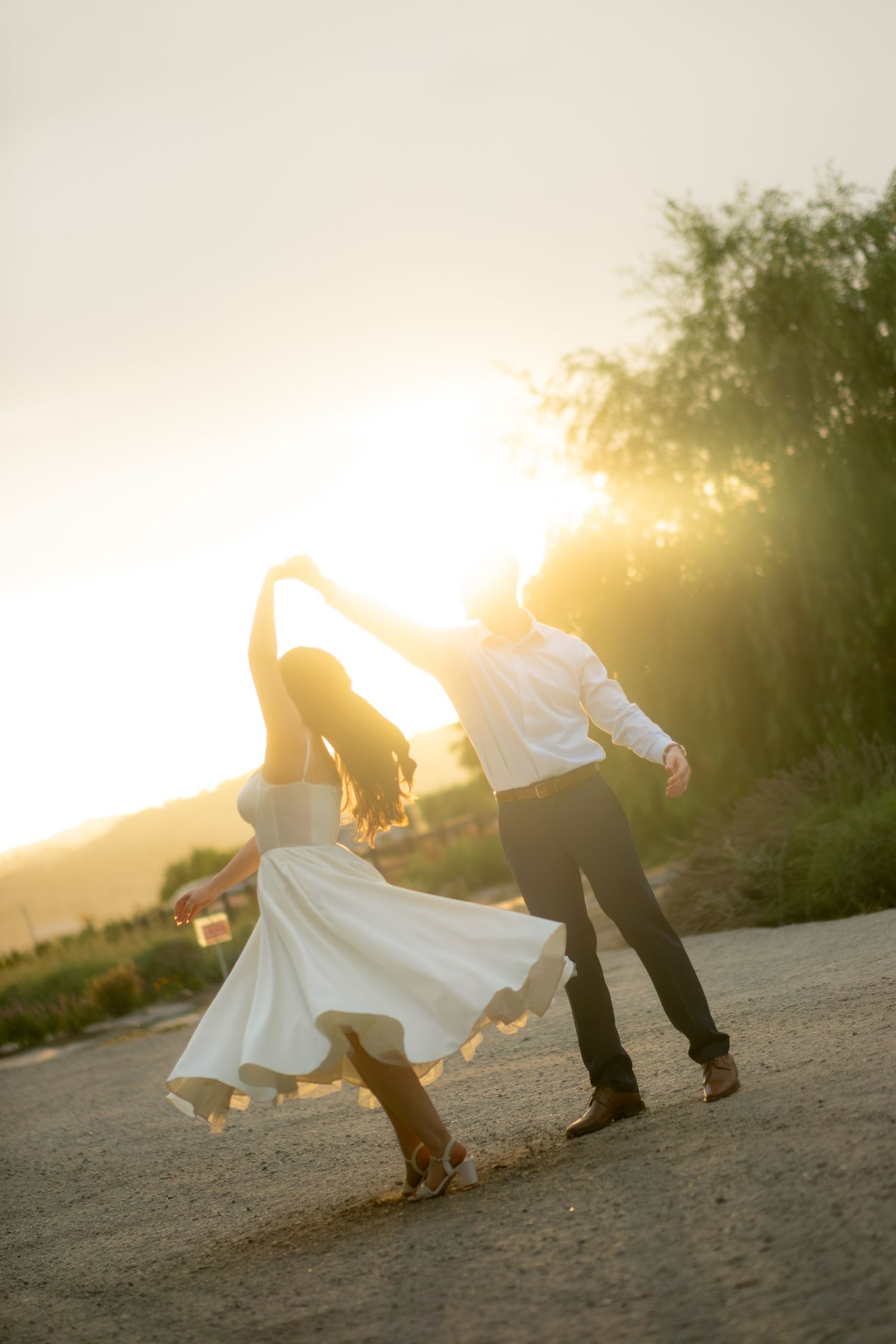A bride in a flowing white dress twirls with her groom on a dirt road at sunset.