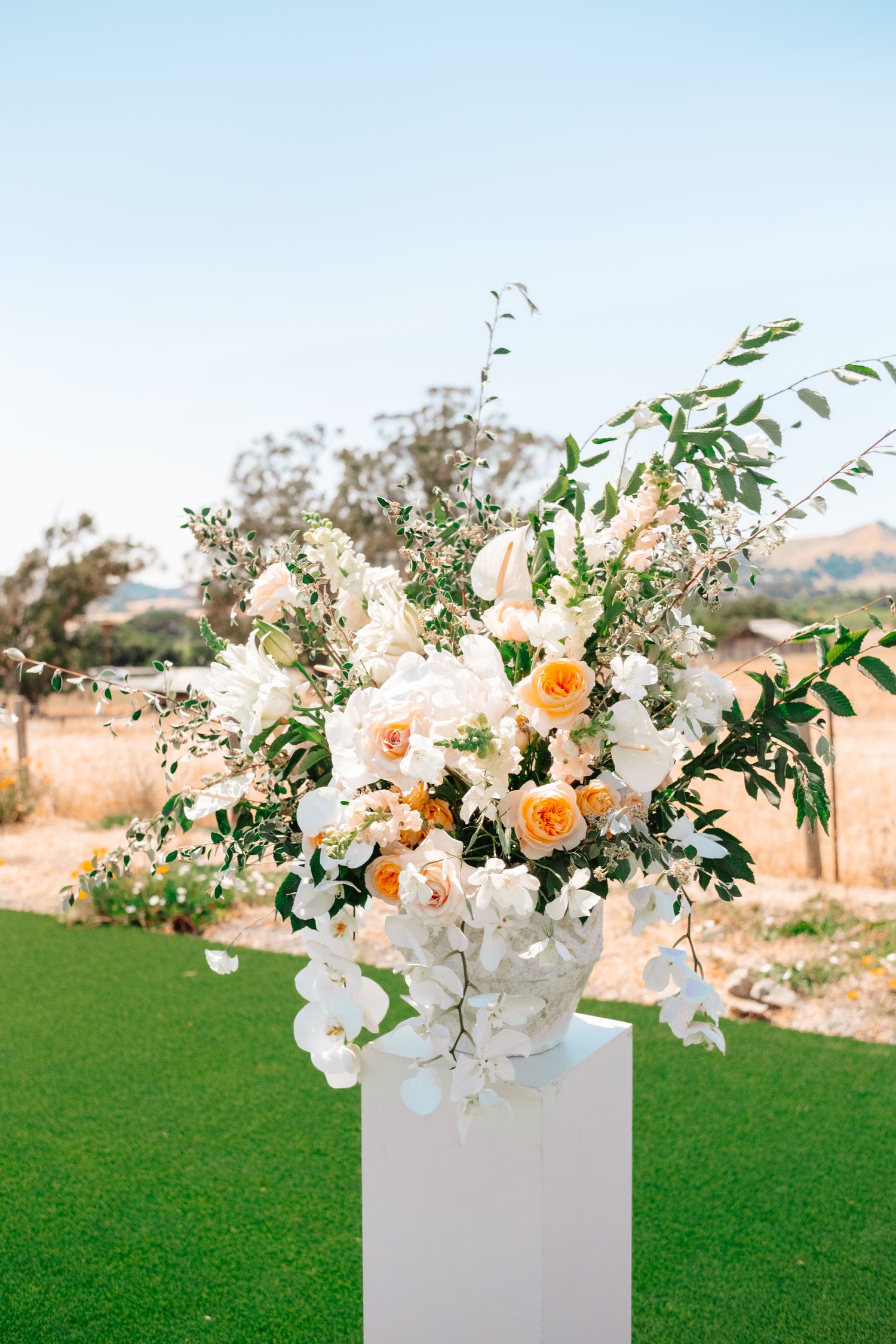 Elegant outdoor wedding floral arrangement with white orchids, peach roses, and greenery on a white pedestal.