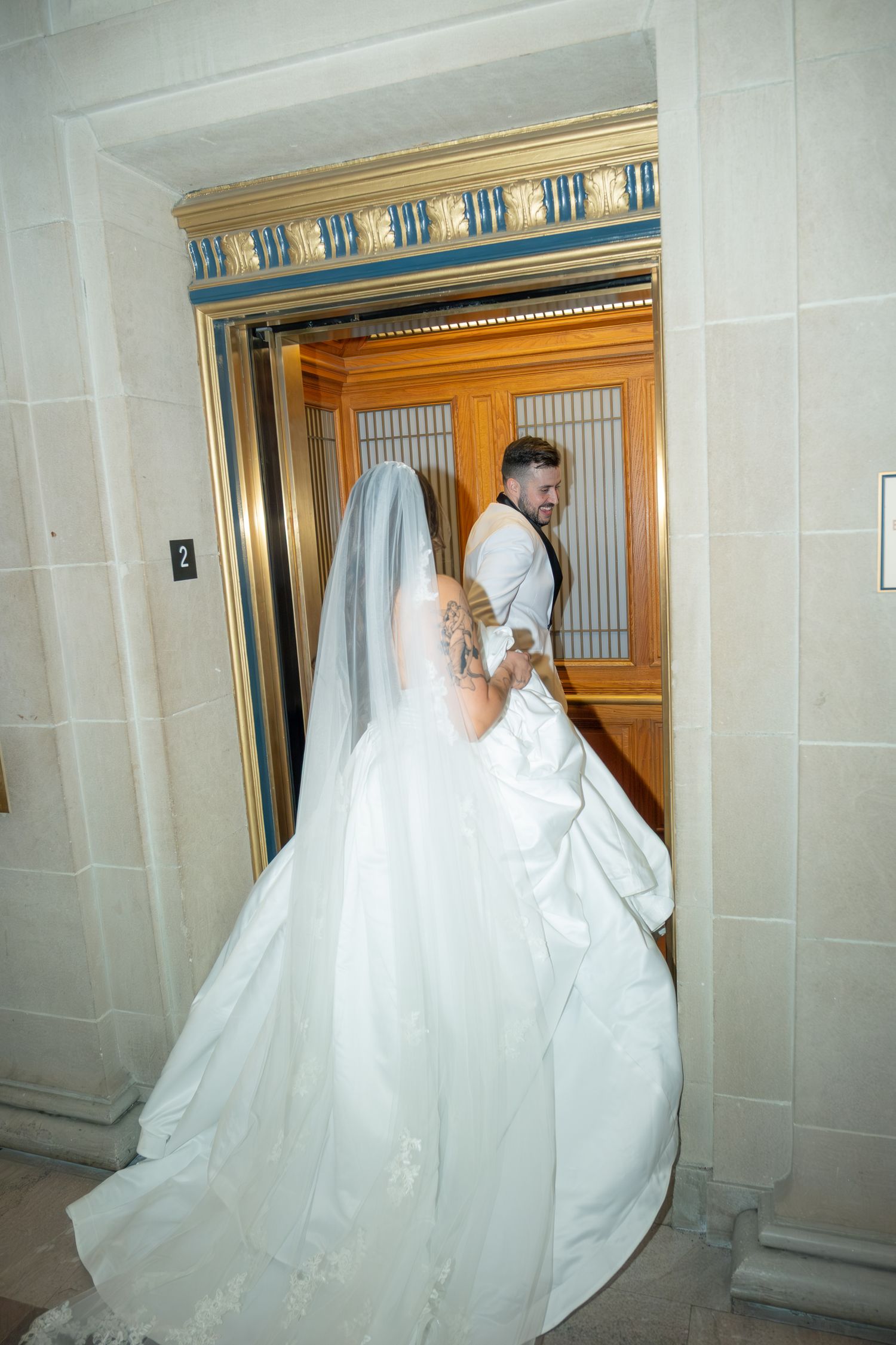 Bride in white gown and veil entering an elegant gold elevator with groom on their wedding day.