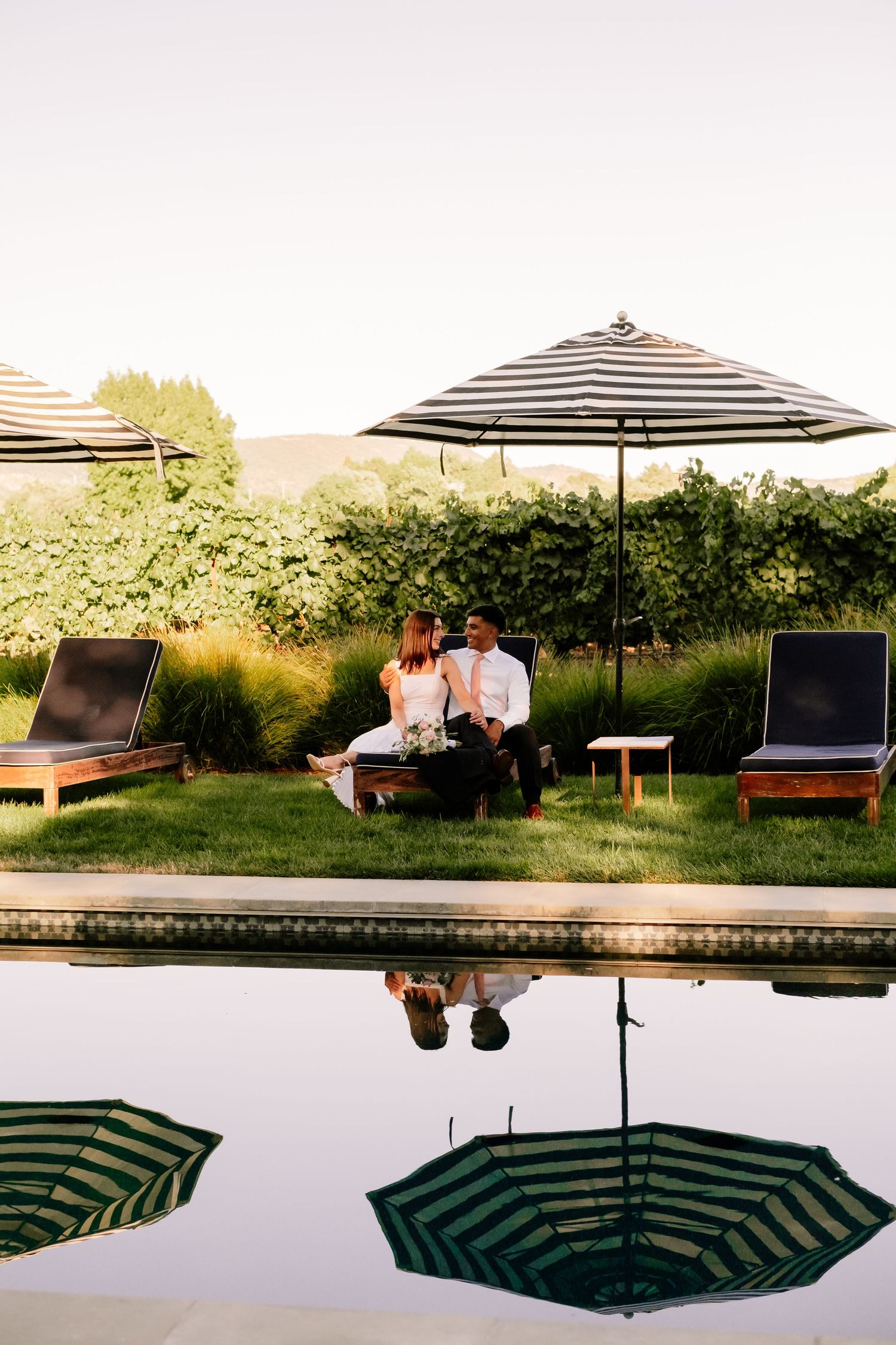 Couple sitting poolside on lounge chairs under striped umbrella with lush vineyard backdrop at sunset.