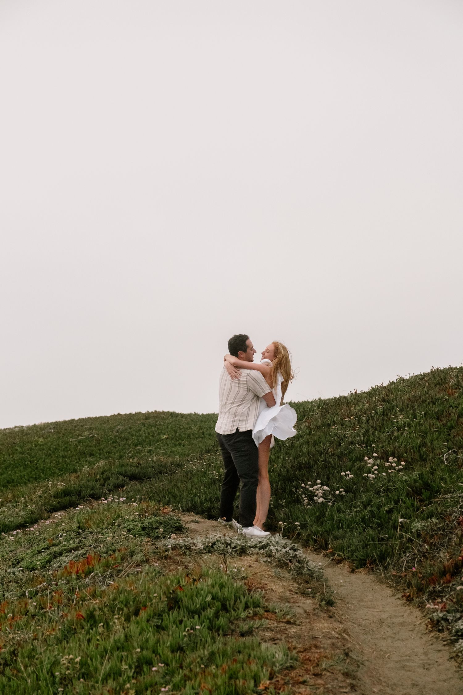 Couple embracing and kissing on a scenic coastal hillside trail surrounded by wildflowers in foggy weather.